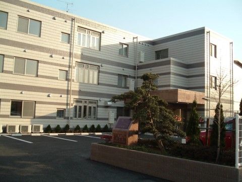 白とベージュが基調の３階建ての建物に、グレーの横ラインが引いてあるデザインになっている。建物の前には駐車場や花壇が作られている。
