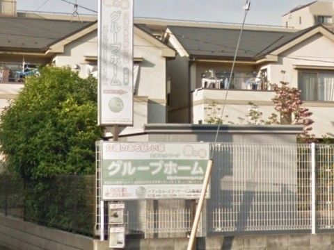 建物前のフェンスに施設名が書かれた２つの看板が設置してある。建物は三角屋根でベランダがあり、壁は白く民家のような外観である。