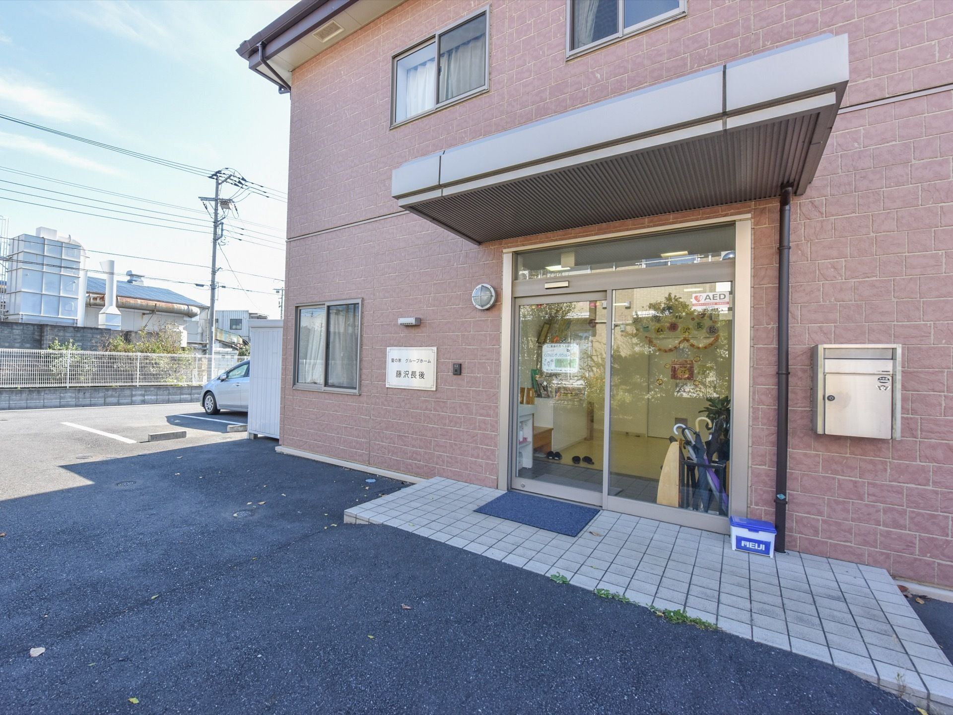 玄関には大きな庇が付いている。壁面には郵便受けと、施設名のパネルがあり、夜間のためにインターホンがある。