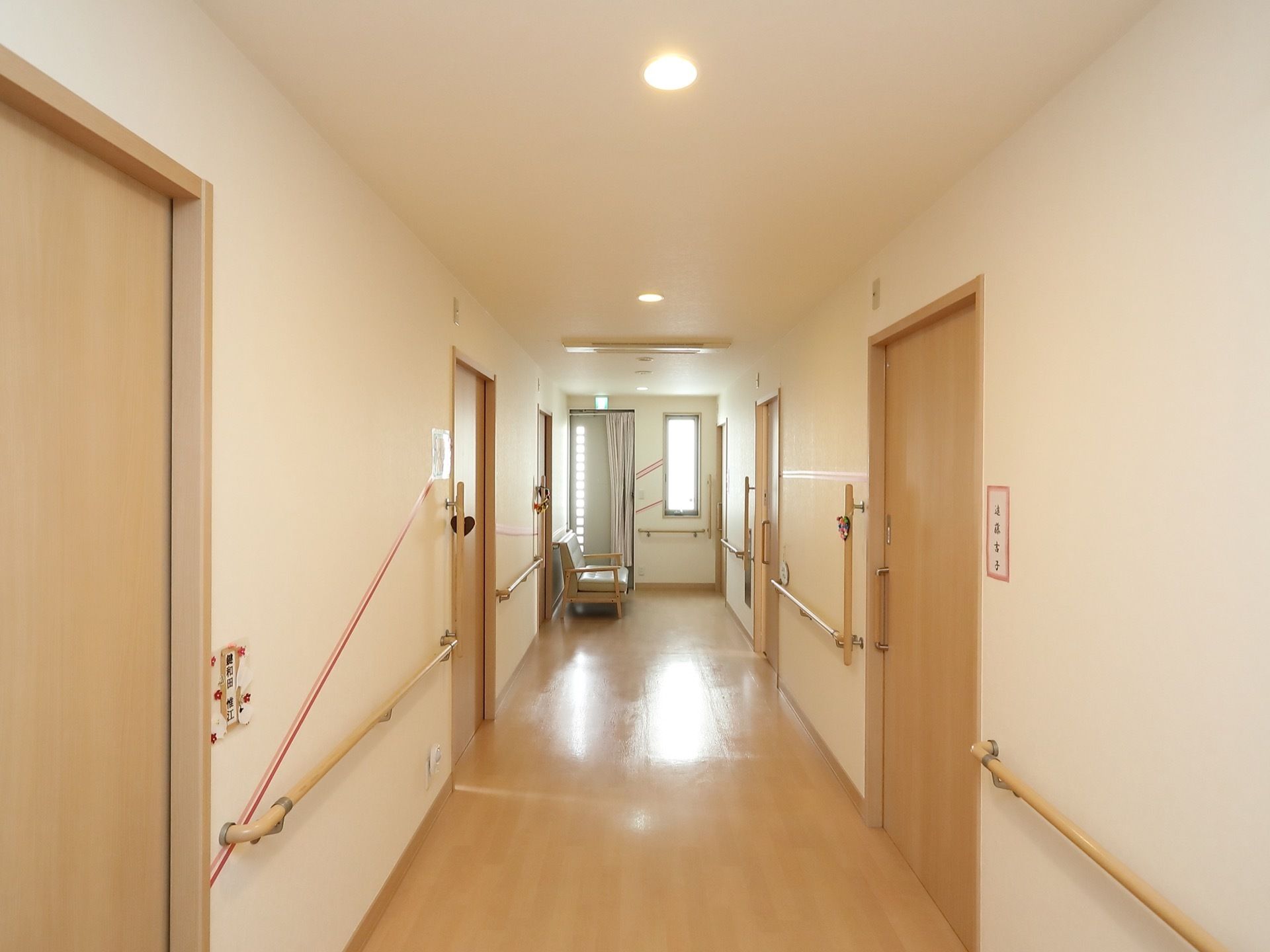 幅が広い廊下は、窓からの採光や照明で明るくなっている。居室入口には入居者様のお名前が表示され、縦方向の手すりが設置されている。