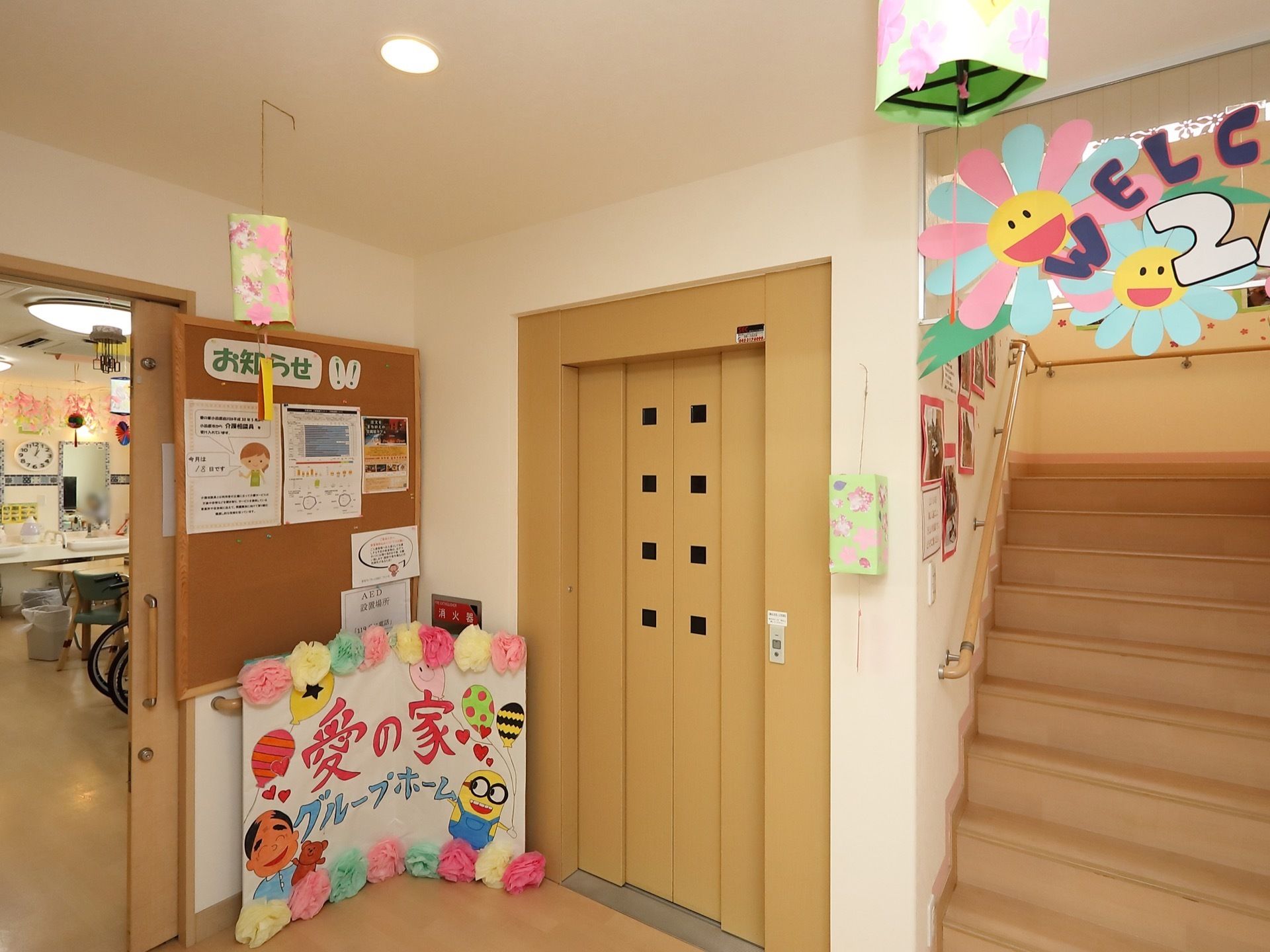 エレベーター乗降口に、楽しい絵や紙で作られた花飾りがついた手作りの看板が置かれている。階段の壁には写真が貼られている。