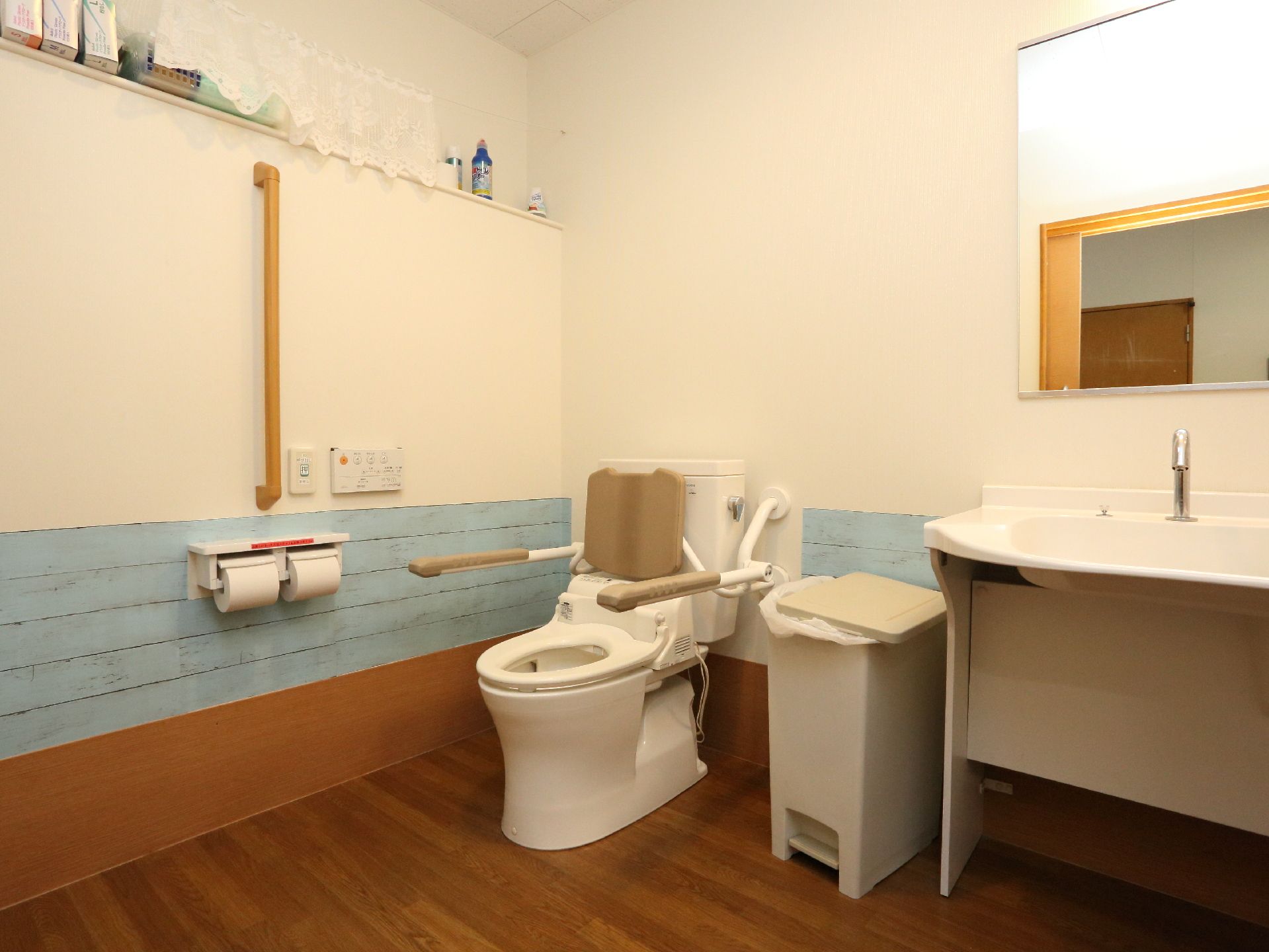 トイレの様子。水洗の洋式トイレが設置され、ゴミ箱が置かれている。便器には可動式の手すりがつけられ、壁にも手すりがつけられている。