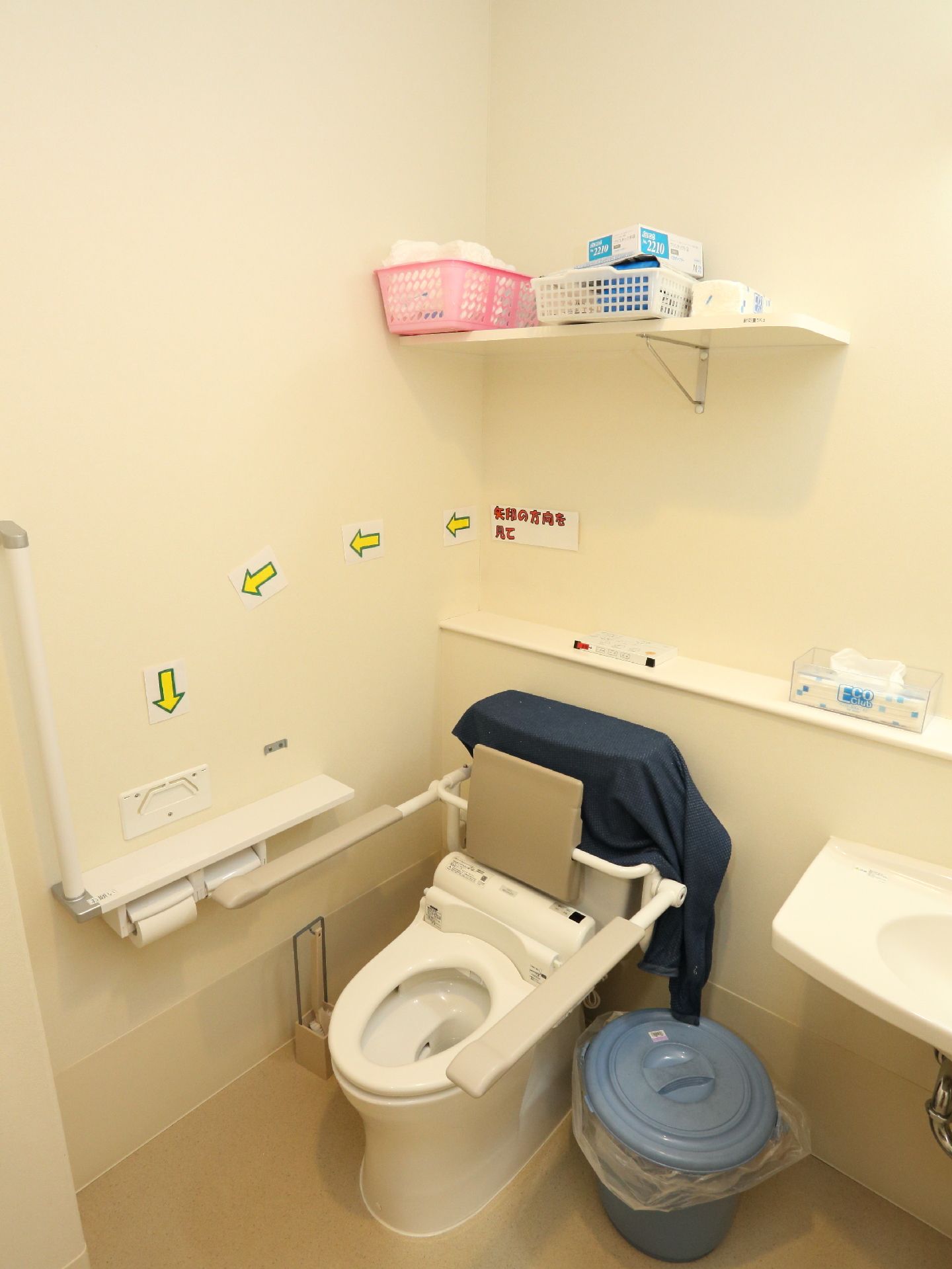 壁の手すりや座面にバリアフリーの設置されたトイレがある。トイレ上部には棚がある。ティッシュペーパーが置かれている。