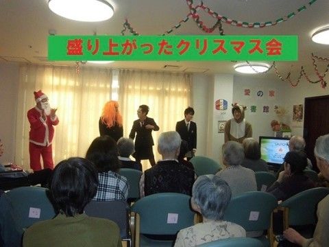 クリスマス会で入居者様が椅子に座り前を見ている。前方ではサンタやトナカイなどに扮したスタッフが立っている。