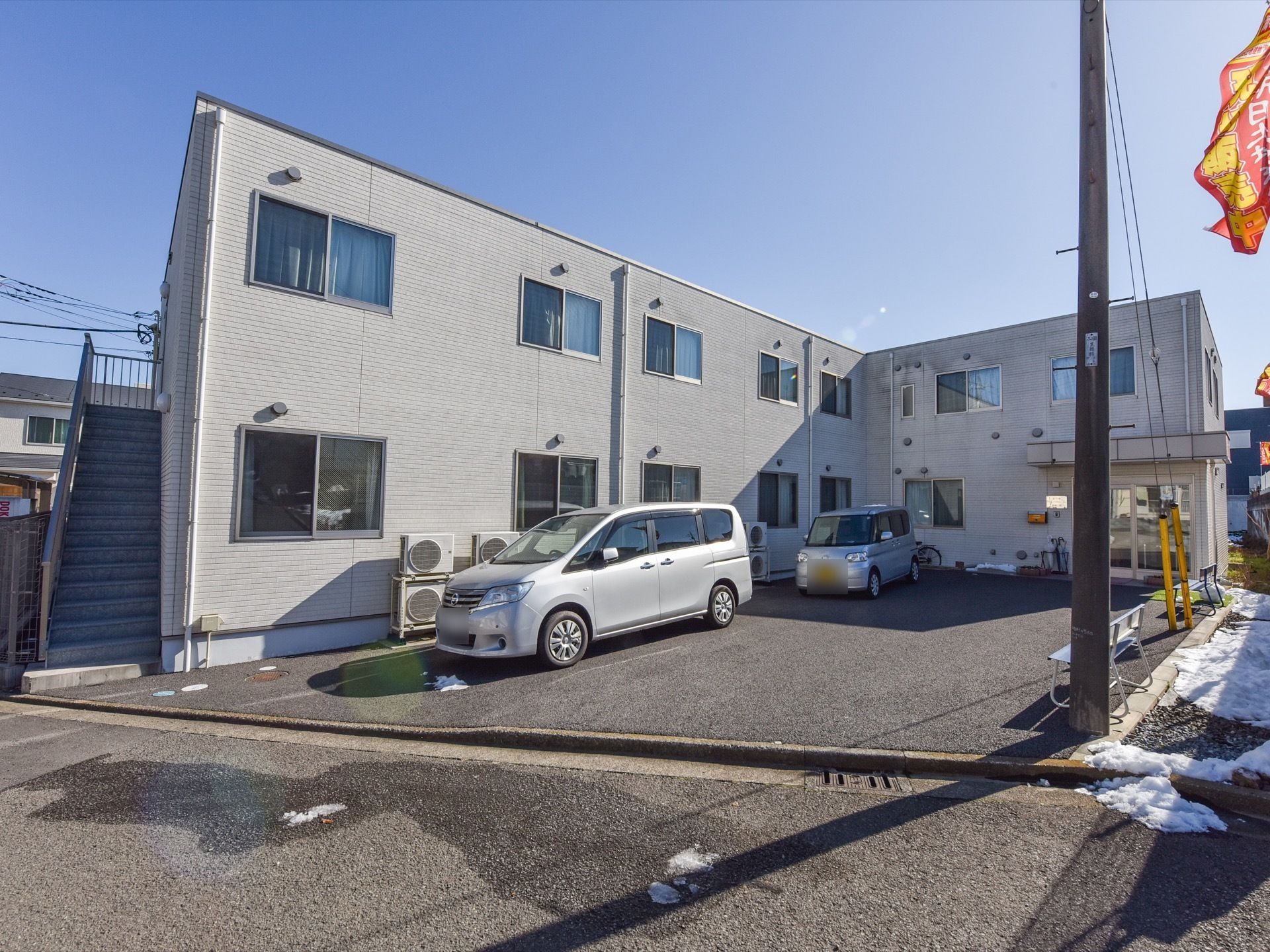 施設の建物は２階建てで、Ｌ字型である。左側に外階段が設置されている。建物の前に、車が２台駐車している。
