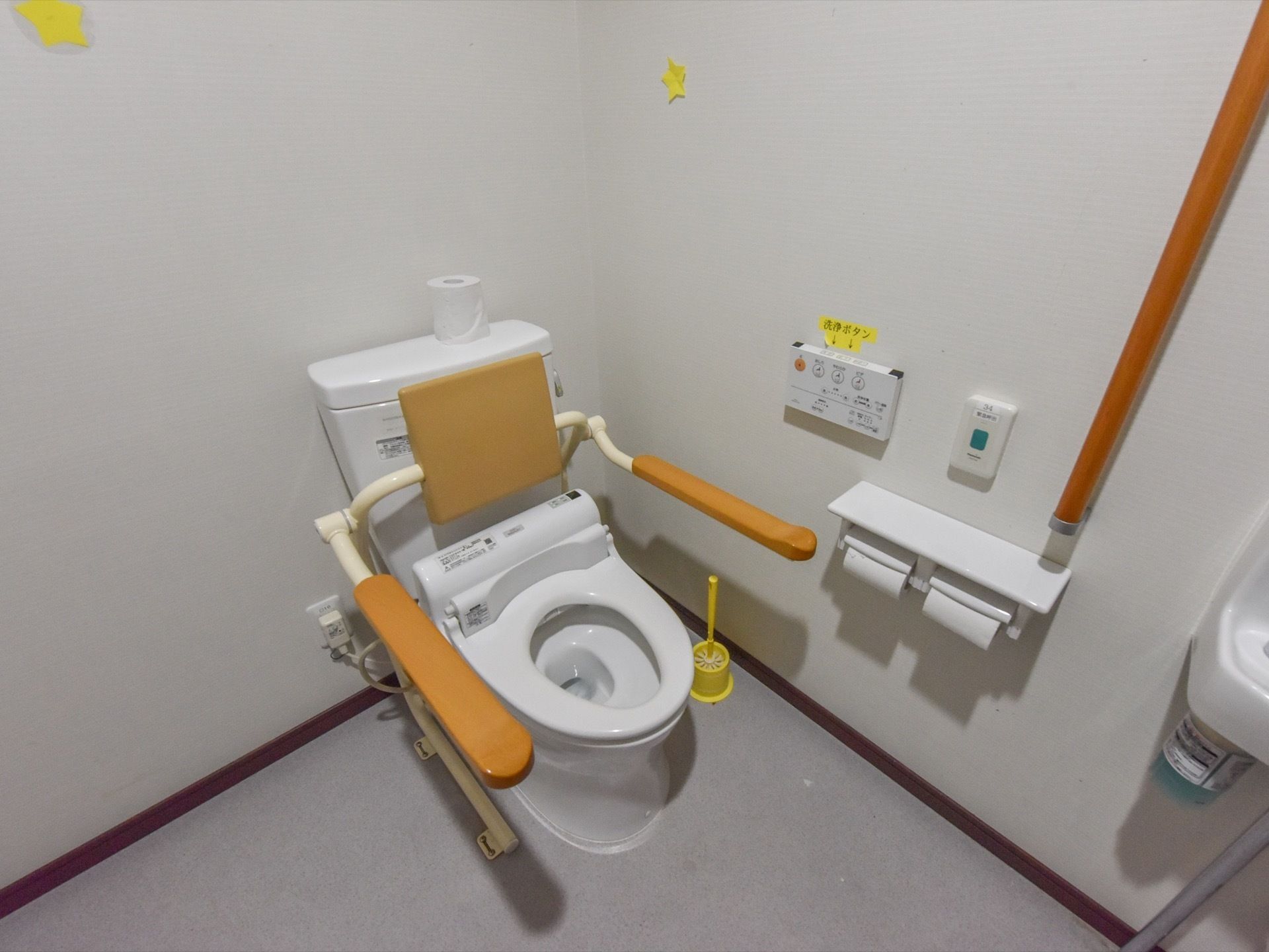 トイレ個室内の様子。温水洗浄便座付きトイレで、壁に操作ボタンがある。便器には、背もたれ付の手すりが設置されている。