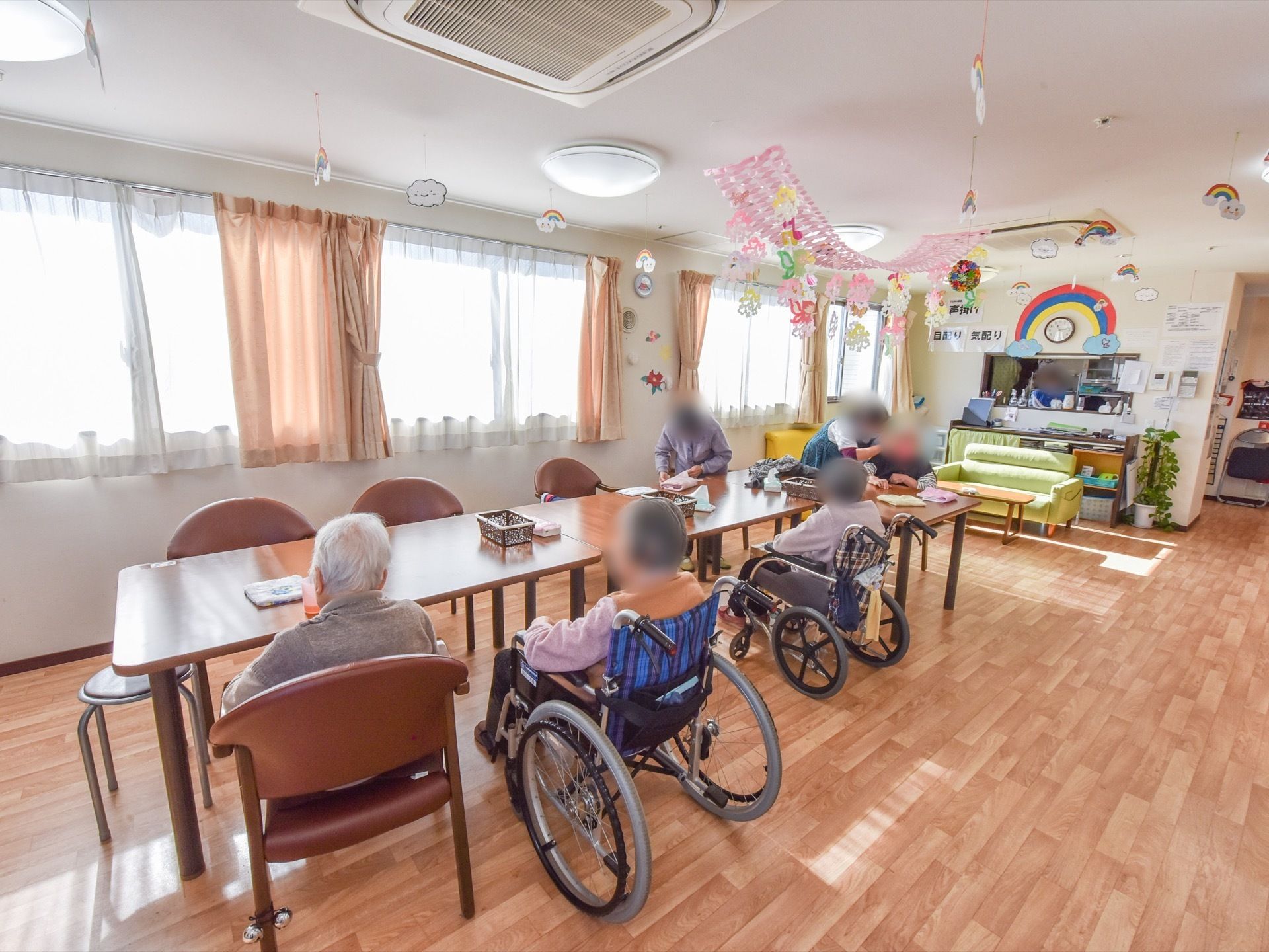 リビングに机が３台置かれており、入居者様たちが椅子や車椅子に座っている。天井には、折り紙などで作られた飾りが吊るされている。