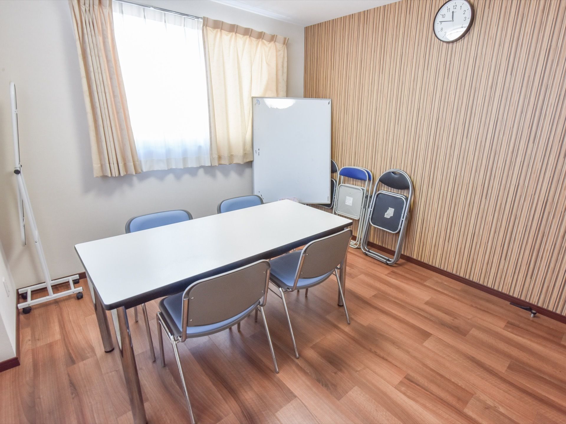 フローリングの部屋に、白いテーブル１台と水色の椅子が４脚ある。壁も茶色で、縦に黒い線の模様が入っている。