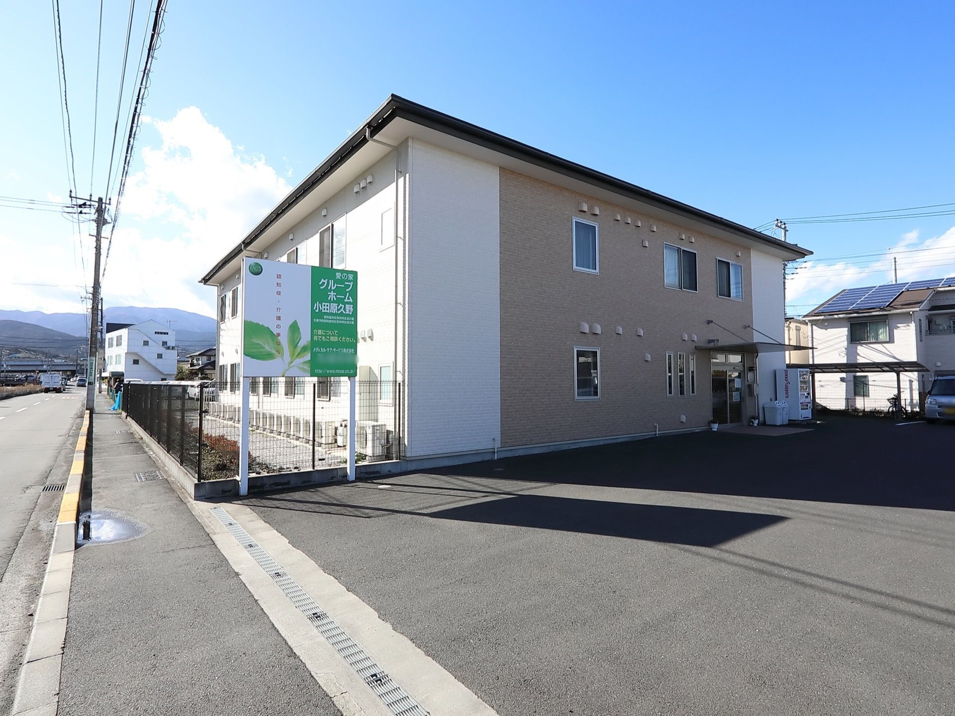 のどかな雰囲気の場所にある施設である。玄関前には、舗装された広いスペースがある。歩道の近くに看板が設置されている。