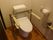 ベージュの壁とブラウンの床の落ち着いた雰囲気のトイレには緊急通報装置が設置されている。便器の脇には可動式の手すりが取り付けられている。