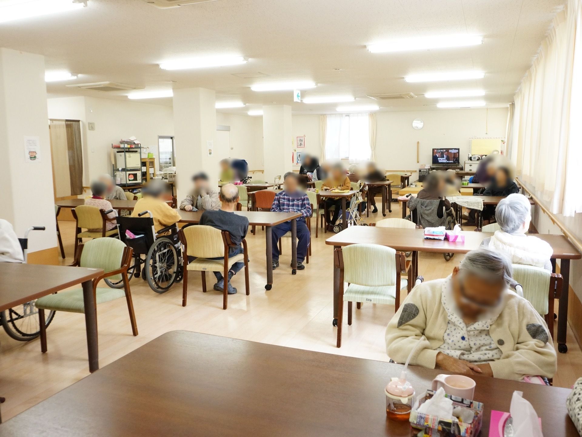 たくさんの入居者様が、テーブルの周りに座ってくつろいでいる。各テーブルの上には、ボックスティッシュが置いてある。