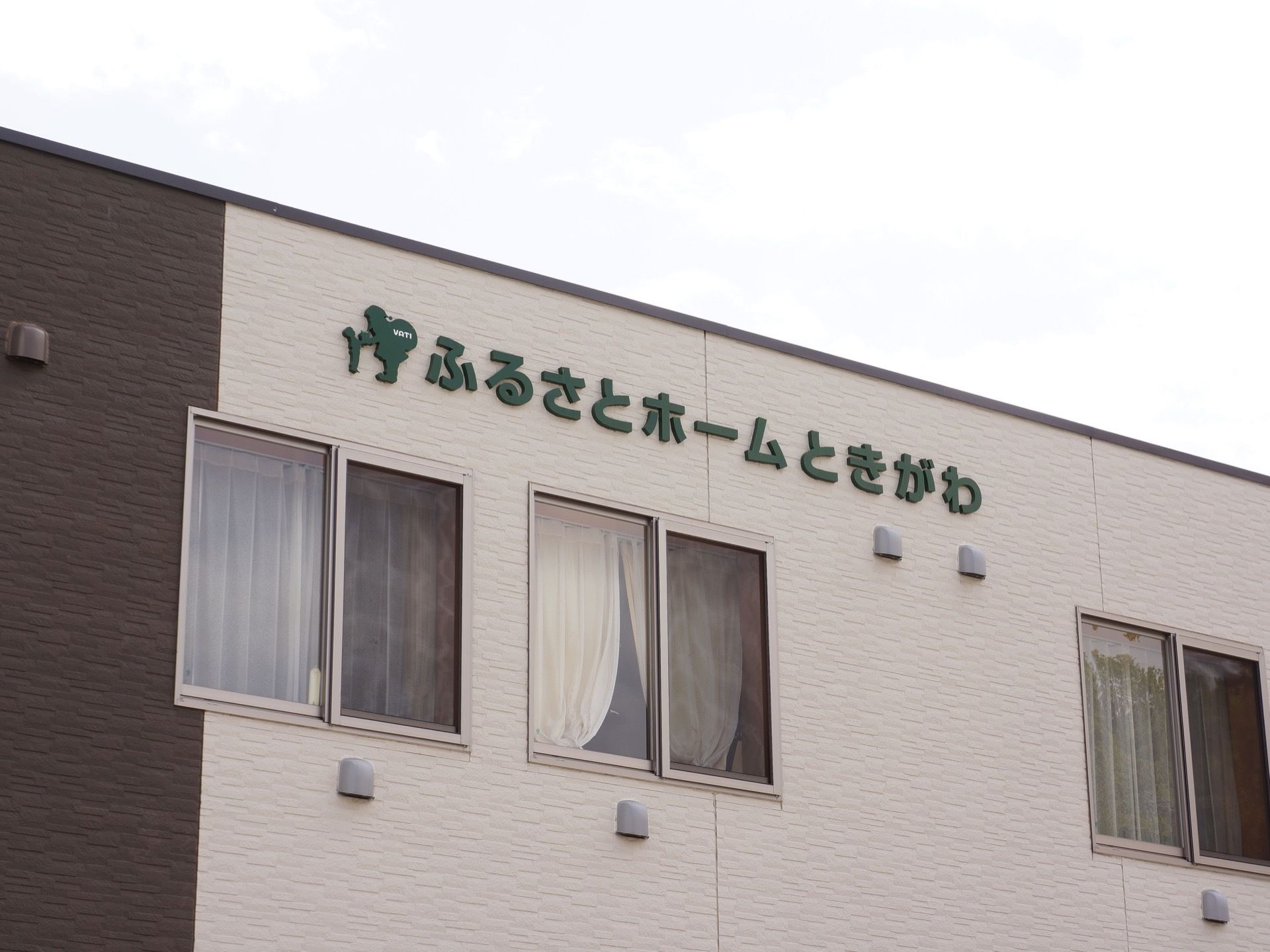 建物の屋根に近い場所に、緑色の大きな文字で施設名が書かれている。その下には窓があり、白いカーテンが掛けてある。