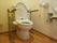 ベージュ色の頑丈な手すりが、トイレの左右に取り付けられている。右側の壁には、オレンジ色の緊急用コールボタンがある。