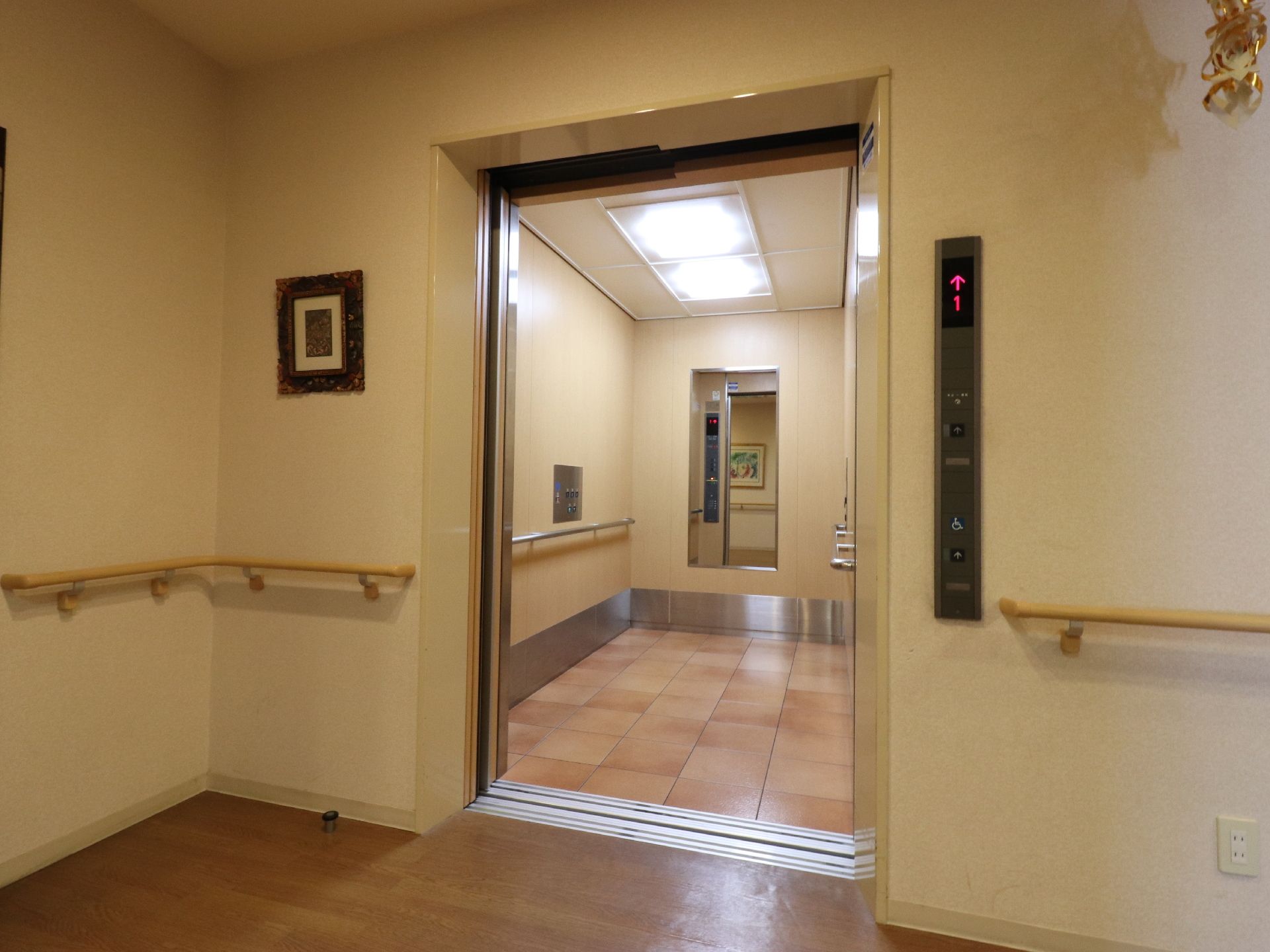 エレベーターの入って正面には、鏡がある。またサイドには、手すりがついている。床には、茶色の正方形が組み合わされている。
