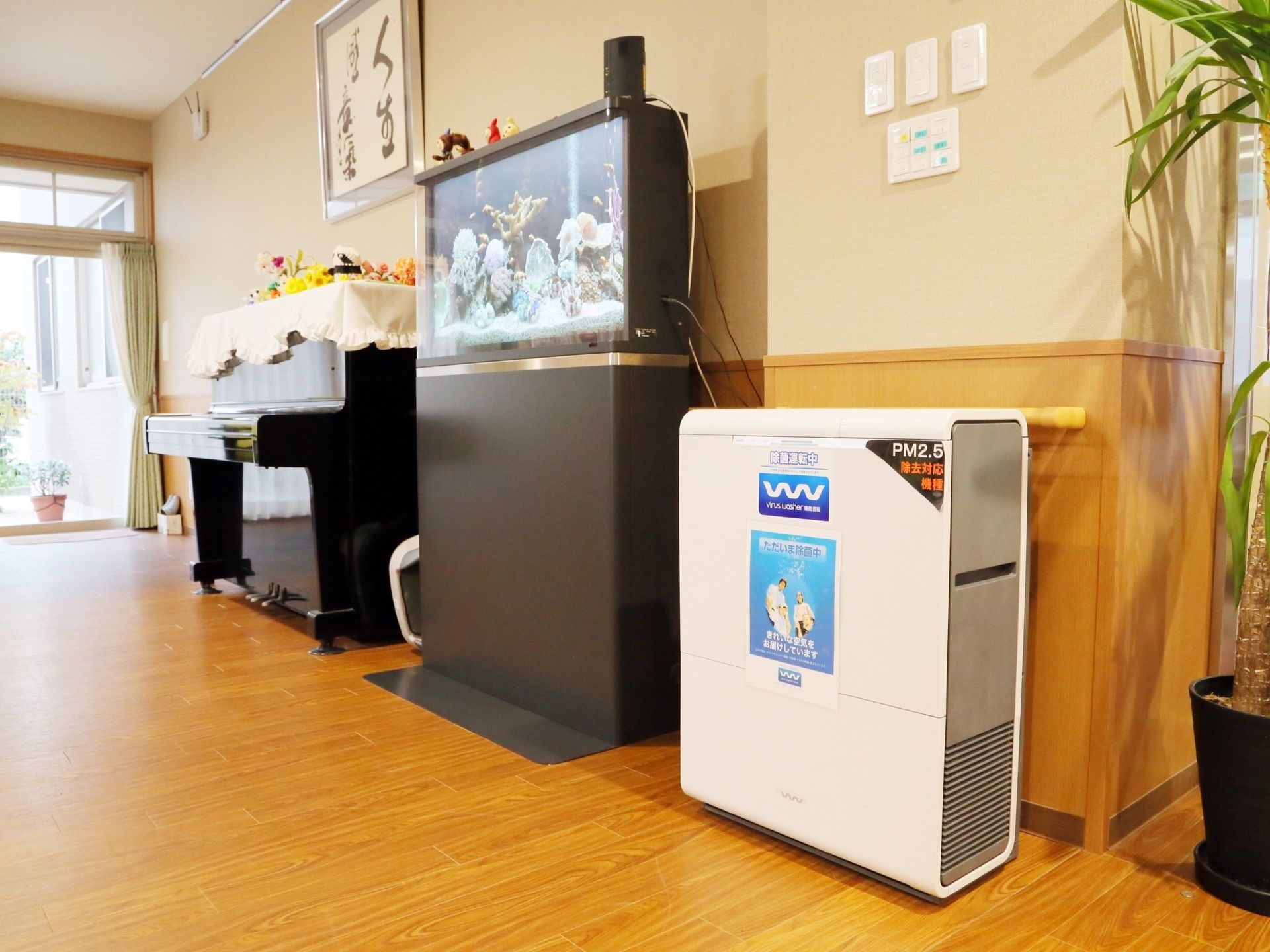 ピアノ、魚の入った水槽が置かれている。空気清浄機と観葉植物がある。壁には文字の描かれた大きな額が飾られている。