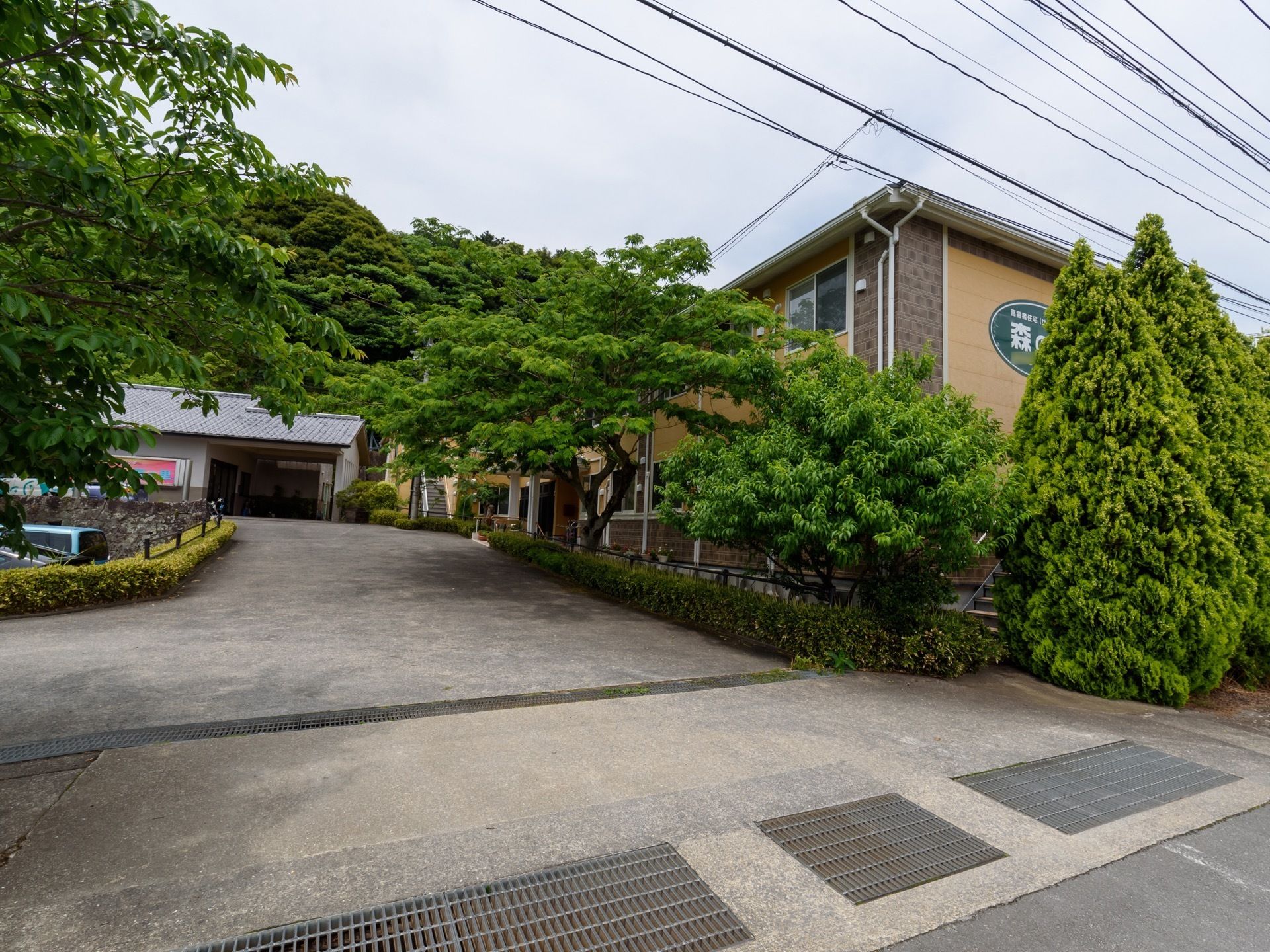 施設の周辺にさまざまな種類の樹々が植えられている。施設はベージュを基調とした、２階建ての建物である。