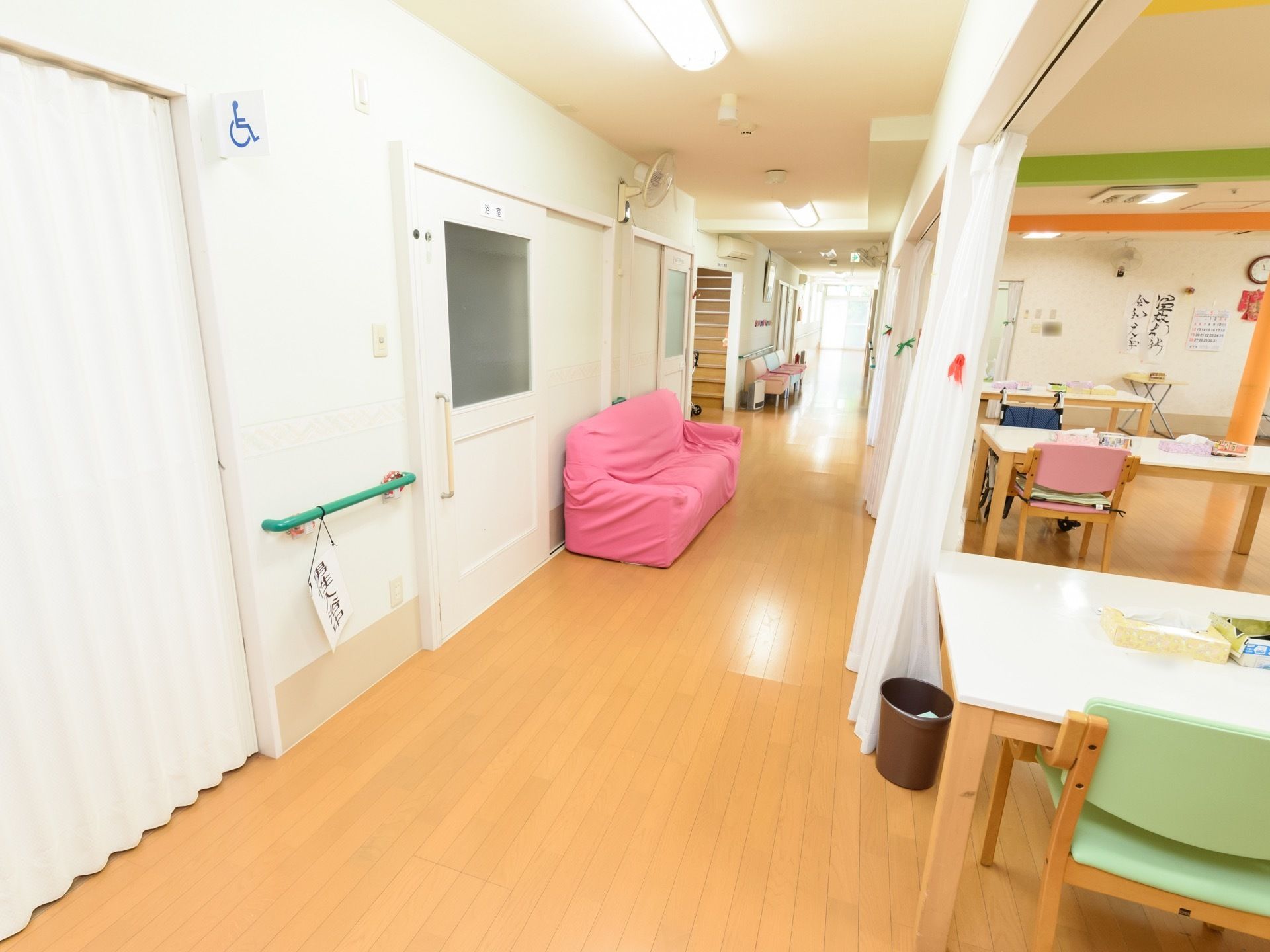 食堂脇の通路にピンクのソファーが置かれている。その奥にはカラフルな椅子が並べられている。居室のドアは白を基調としている。