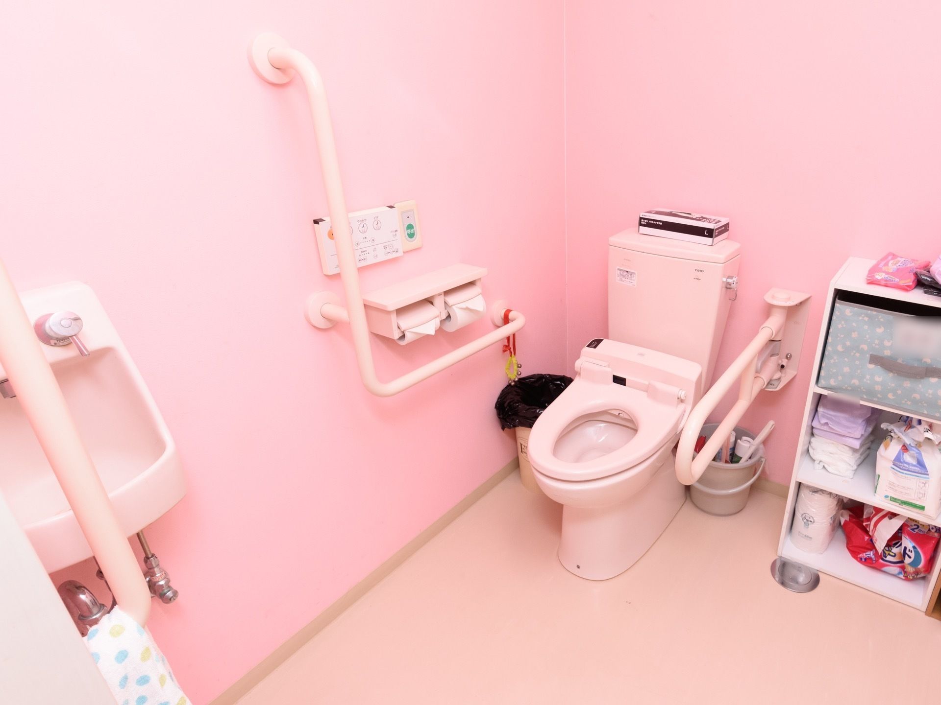 薄いピンク色で統一された個室トイレである。便座の横に折り畳み式、固定式の手すりが取りつけられている。