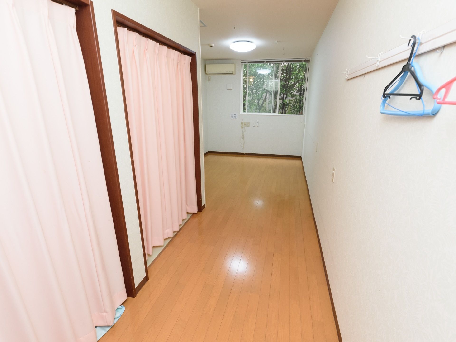 フローリングの床、白い壁の居室である。左手にピンクのアコーディオンカーテンで仕切られたスペースが２か所ある。