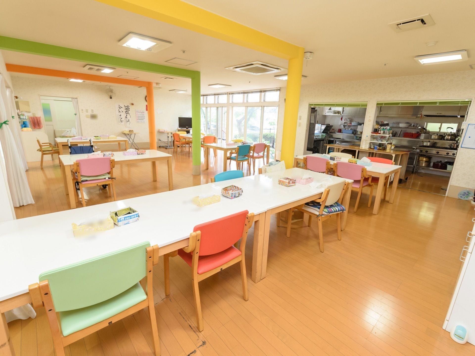 大きな白いテーブルに緑やオレンジ、ピンクなどの椅子が並べられている。右奥にキッチンがあり、その横に大きな窓がある。