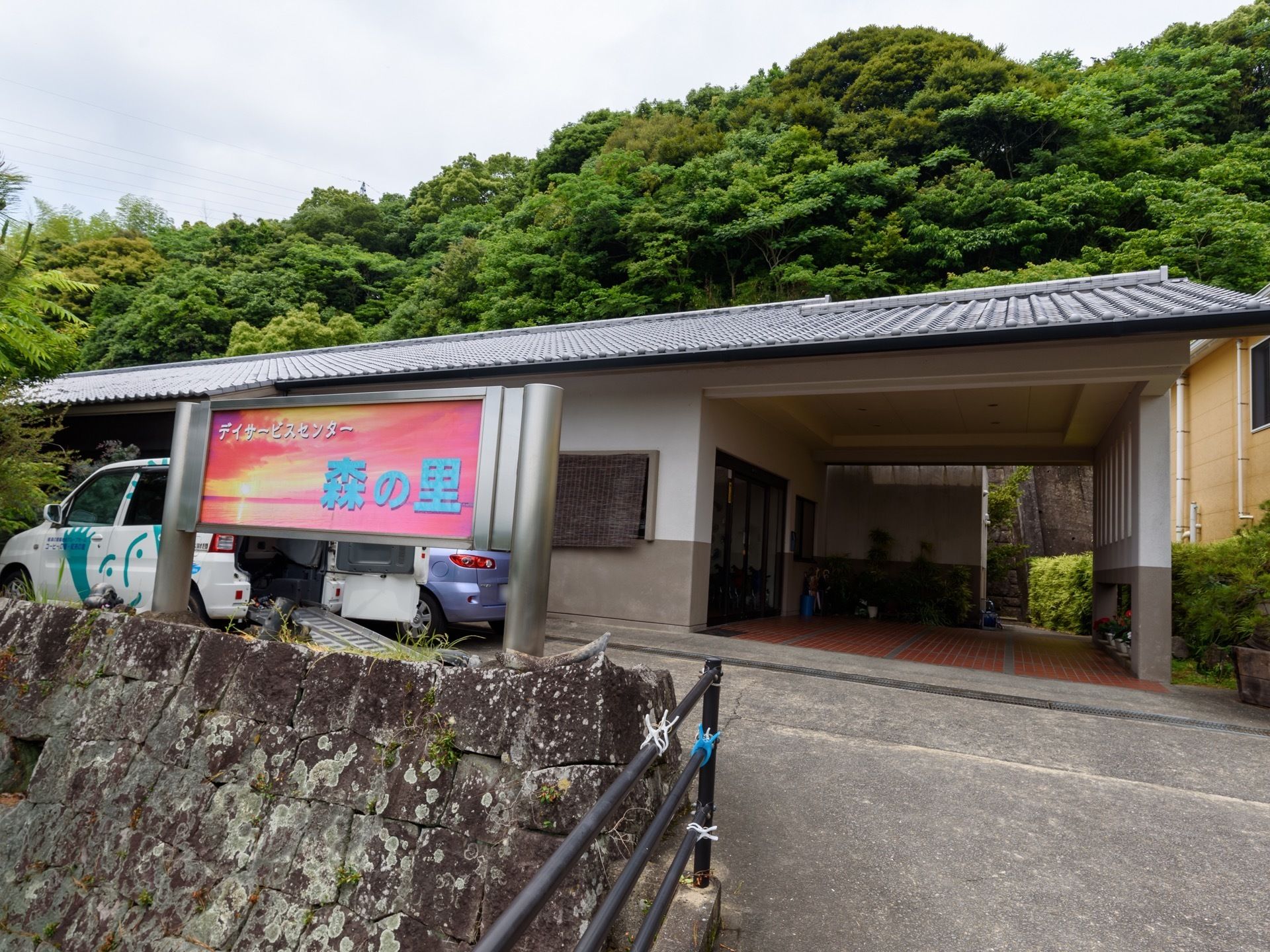 石垣が積まれた上に施設が立っている。玄関ポーチは瓦屋根になっている。建物の前に車が２台停まっている。