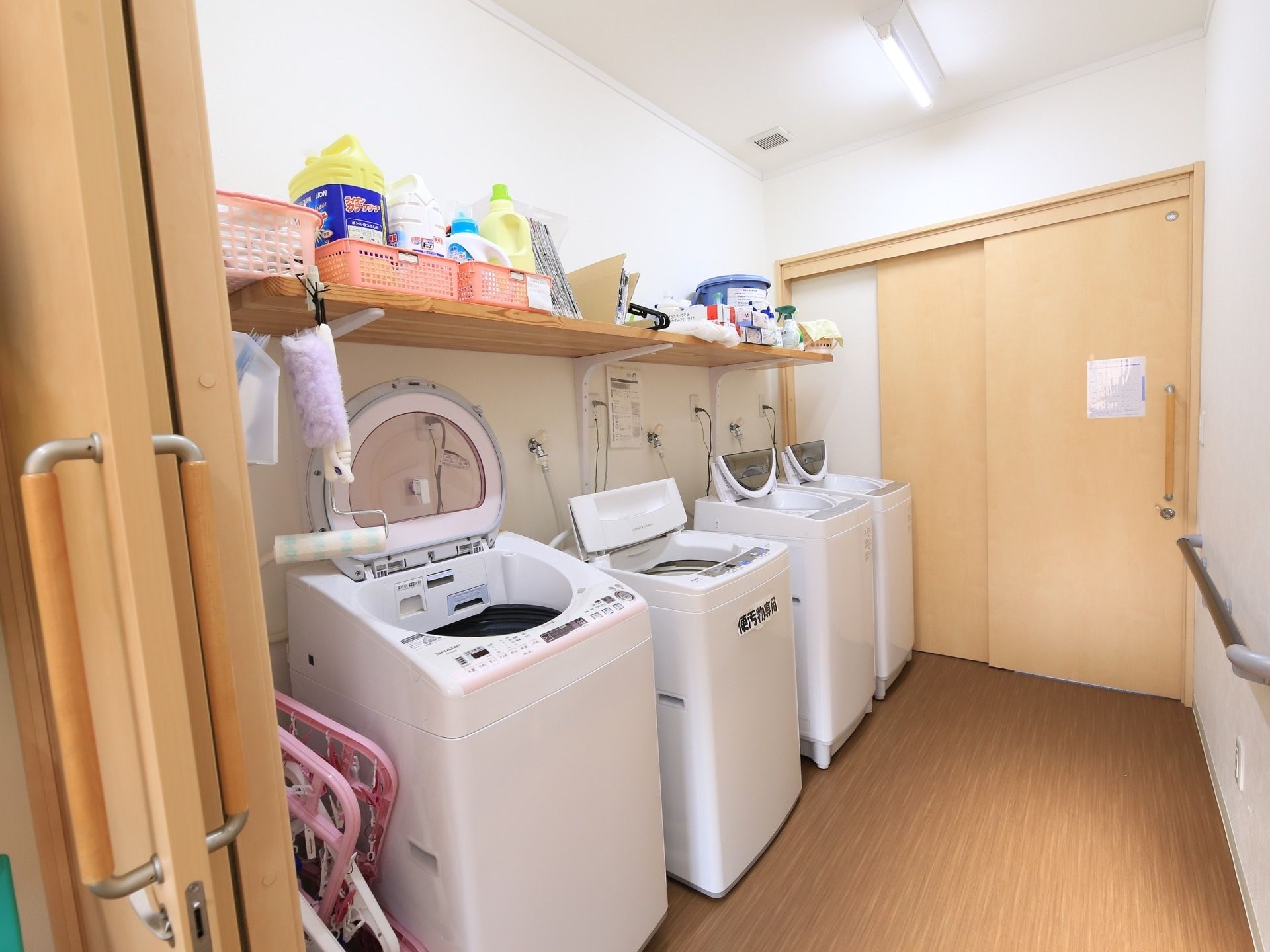 白を基調とした清潔感のあるスペースである。全自動洗濯機が４台設置されている。棚の上には洗剤が置かれている。