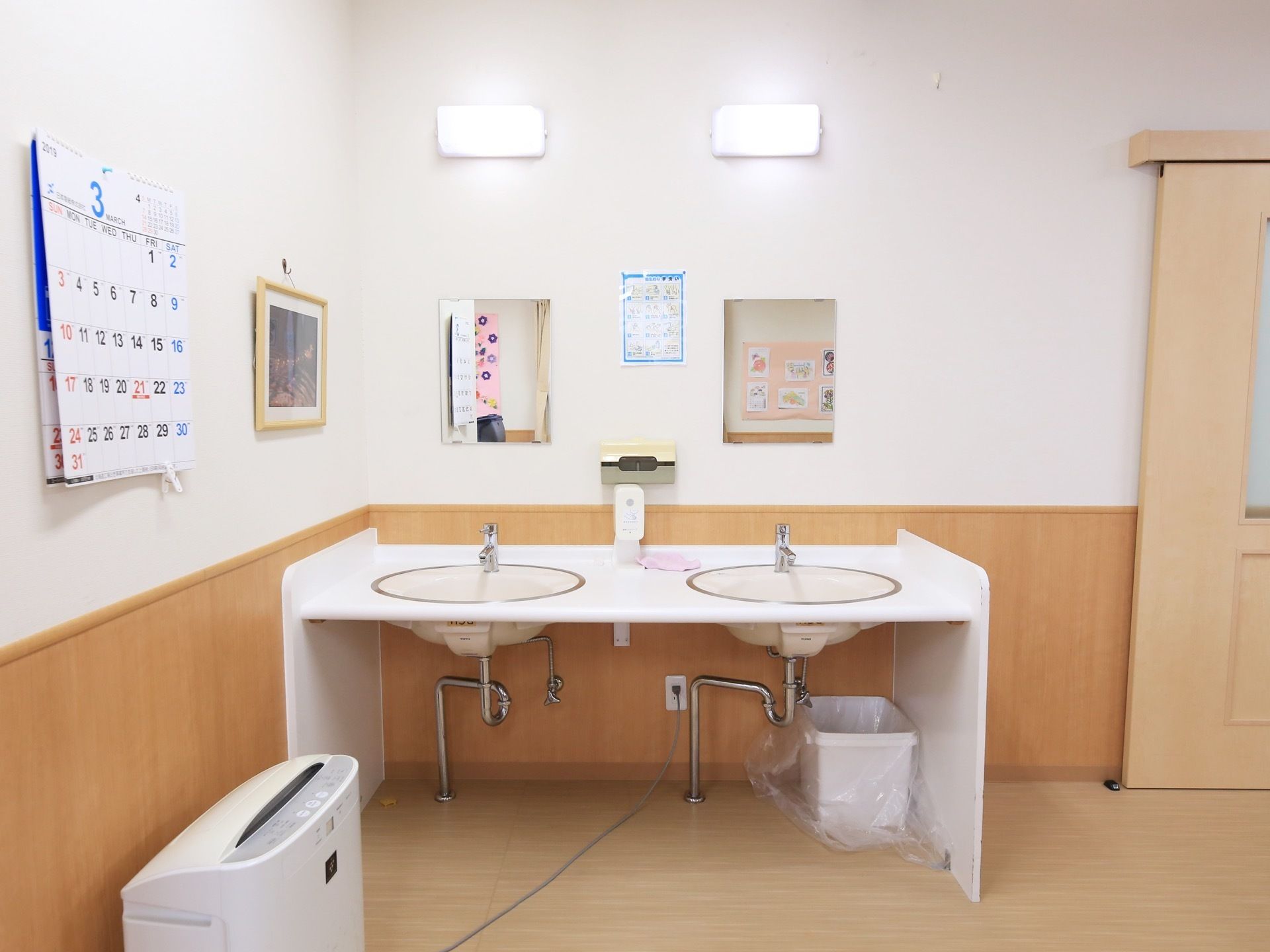 部屋の隅に、２つの洗面台が連結して設置されている。壁面には照明と鏡が取りつけられており、足元にごみ箱がある。