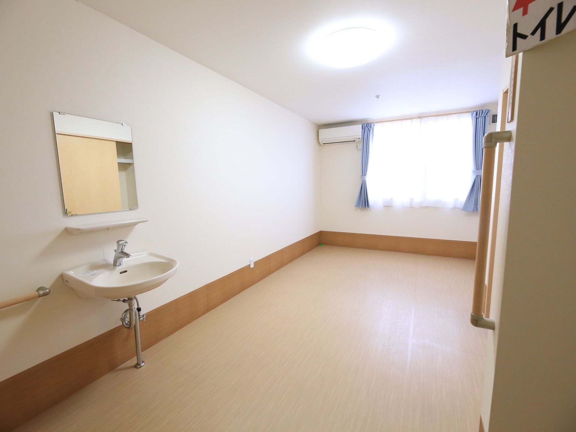 部屋の奥の壁面に窓が施されており、青いカーテンが取りつけられている。左側に洗面台と鏡があり、右上にトイレの案内表示が見える。