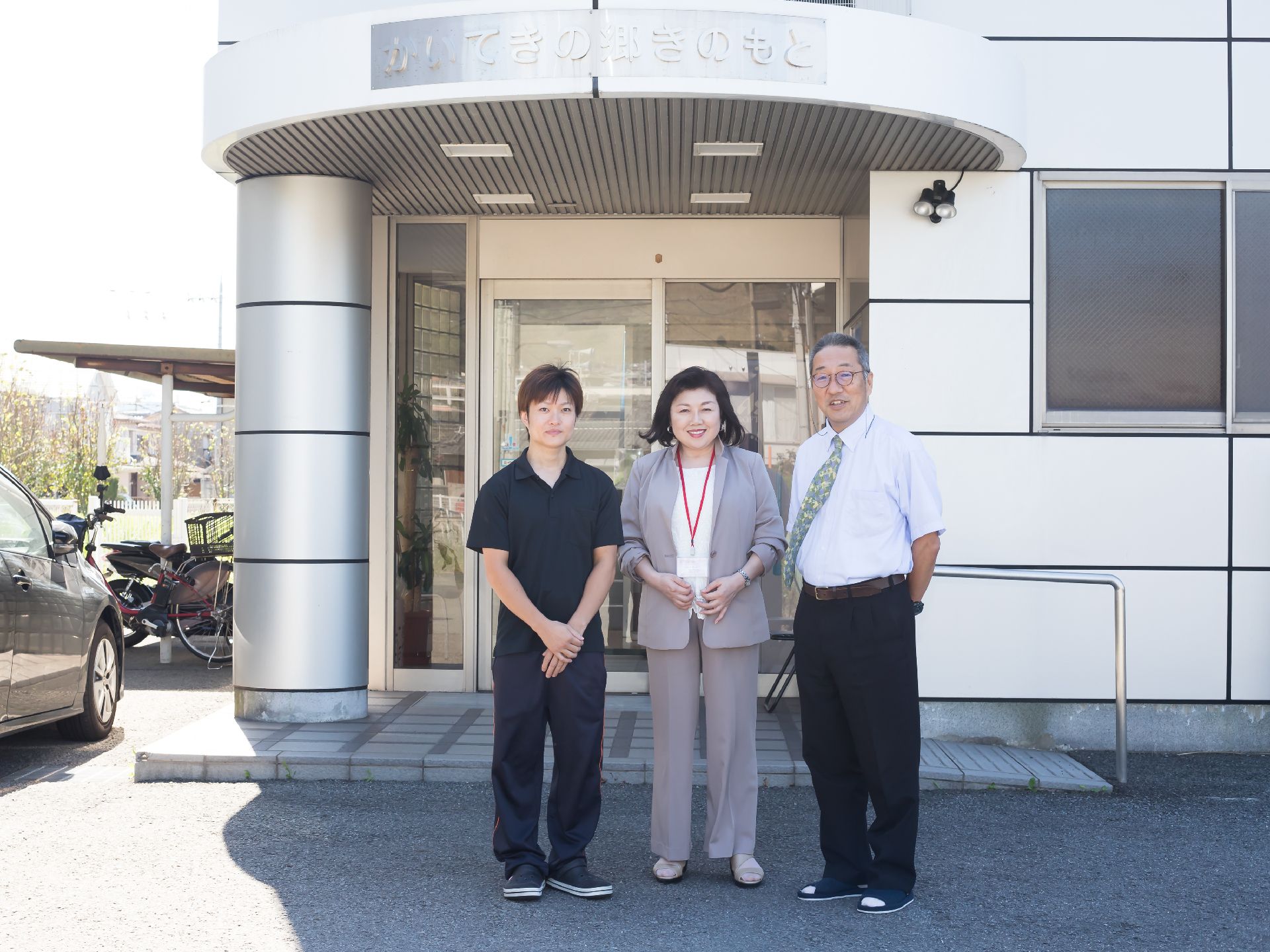 施設名が大きく書かれた建物の玄関前で微笑む３名のスタッフ
