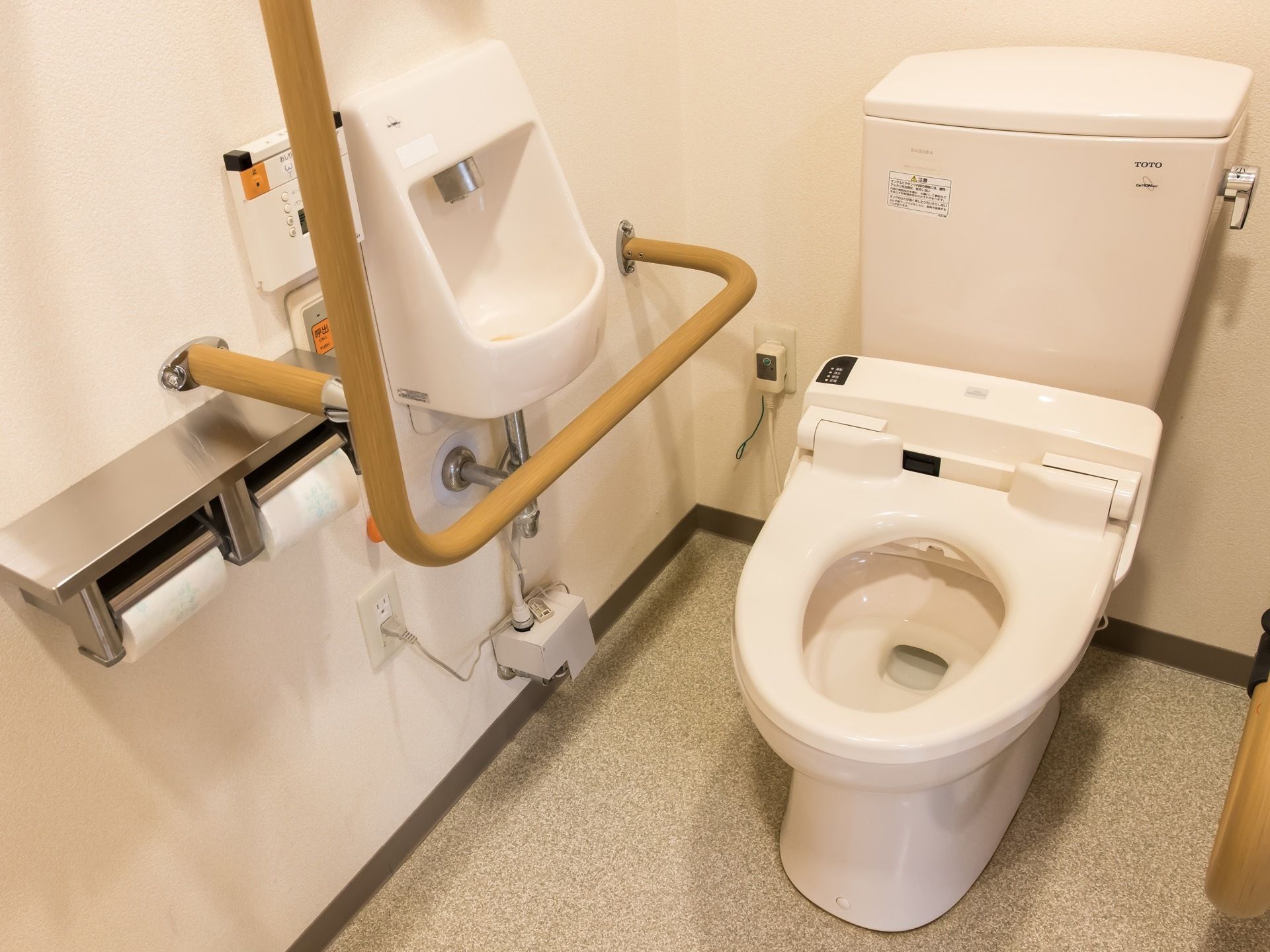 トイレットペーパーホルダーのすぐ側に、緊急通報装置や手すり、手洗い場が取り付けられている。手すりの側には、コンセントがある。