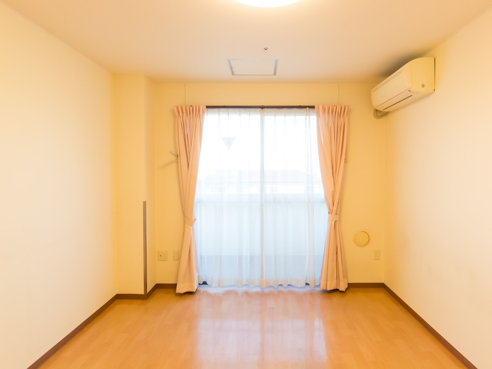 窓には、レースカーテンとピンク色のカーテンが付いている。窓の外には、ベランダがある。窓の側には、エアコンが付いている。