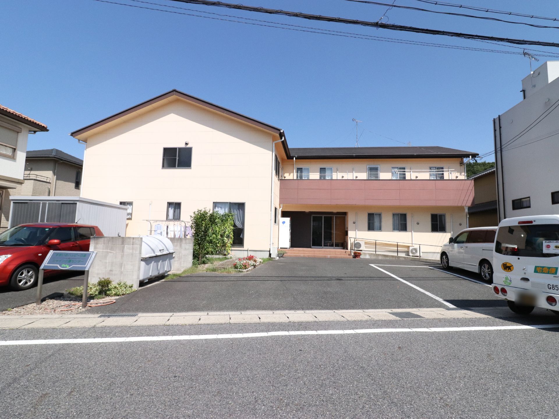 ツートンカラーの2階建ての建物には、玄関前に低い階段とスロープが設けられている。道路に面した場所に施設名の書かれた看板がある。
