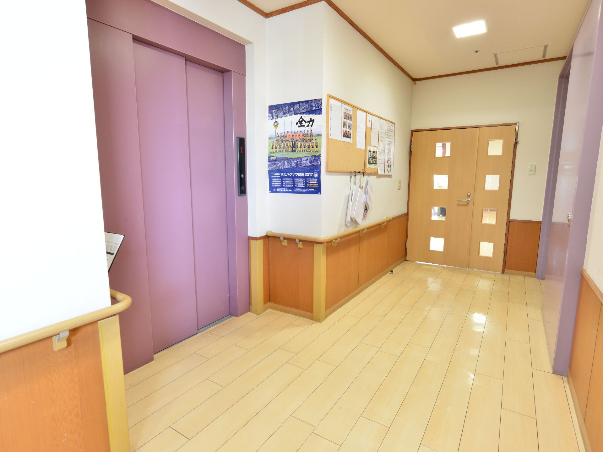 施設のエレベーターホールにはピンク色のエレベーターが設置されている。ホールの壁にはポスターや掲示板などが貼られている。