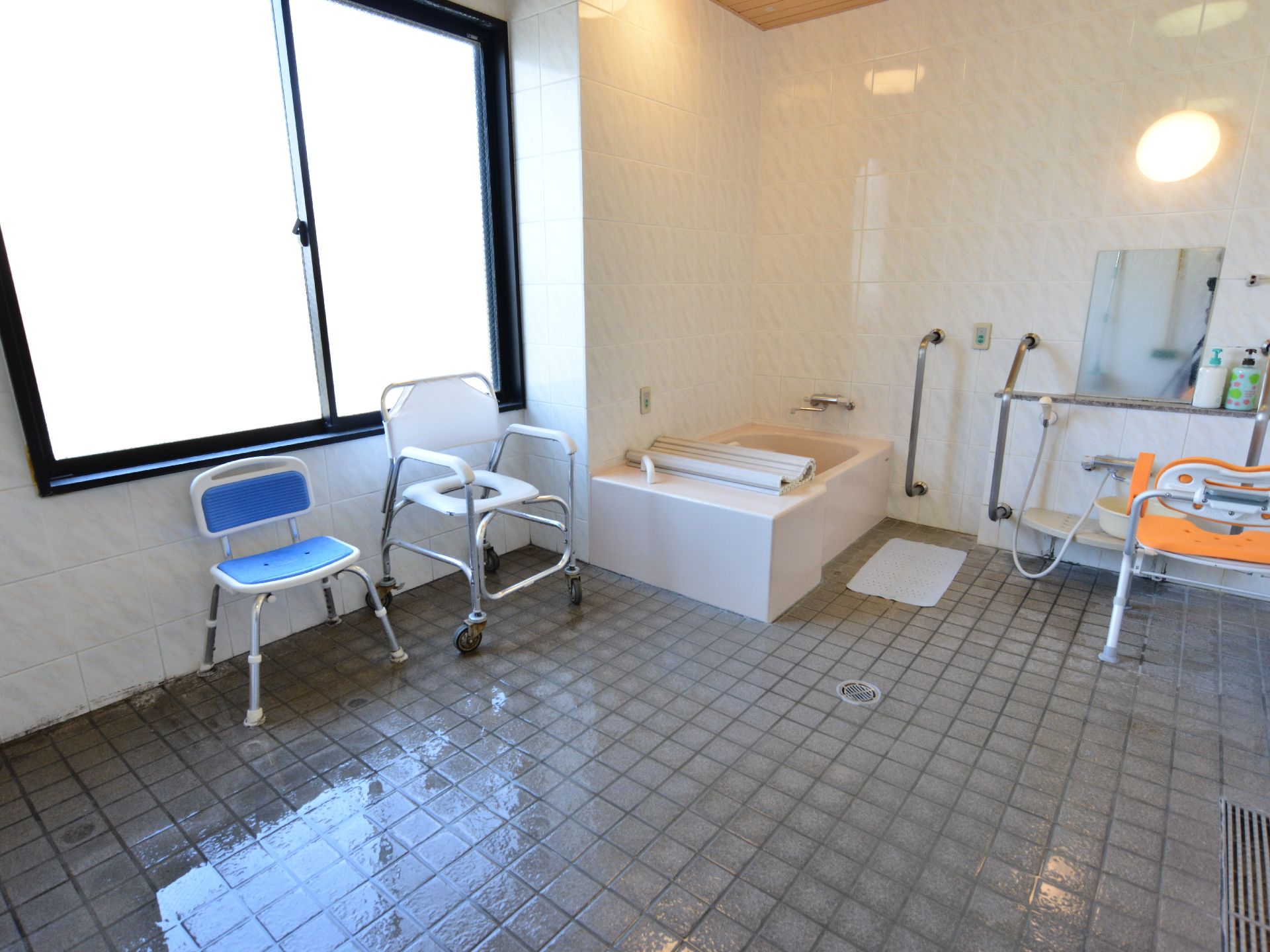 施設の浴室は白壁にタイル張りの床。隅に1人用の浴槽が設置され壁にはシャワーもある。近くには手すりがあり浴室用の椅子も置かれている。