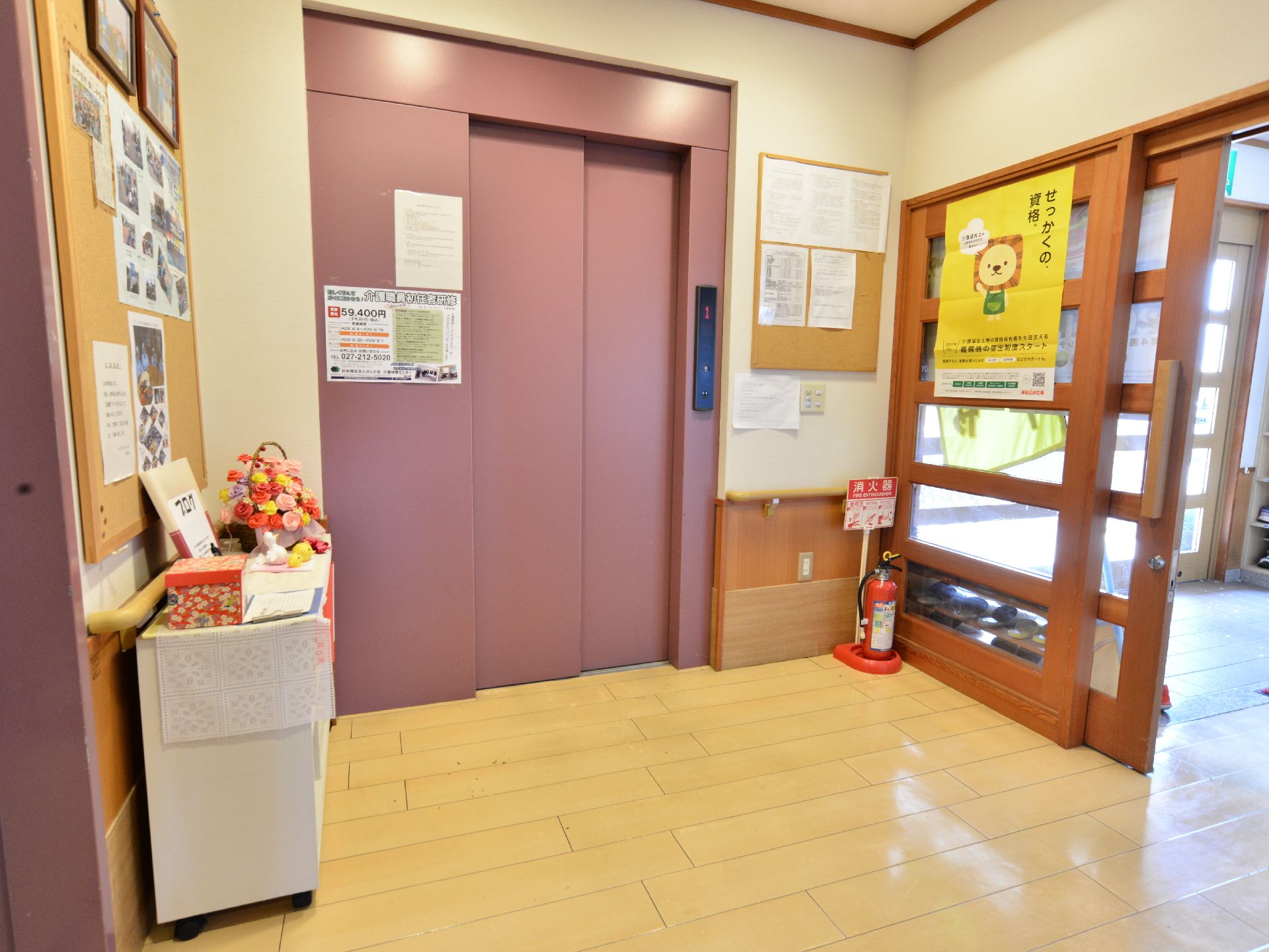 施設の玄関の横の扉の先にはピンクのエレベーターが設置されている。ホールには消火器や掲示板、ポスターや写真などが貼られている。