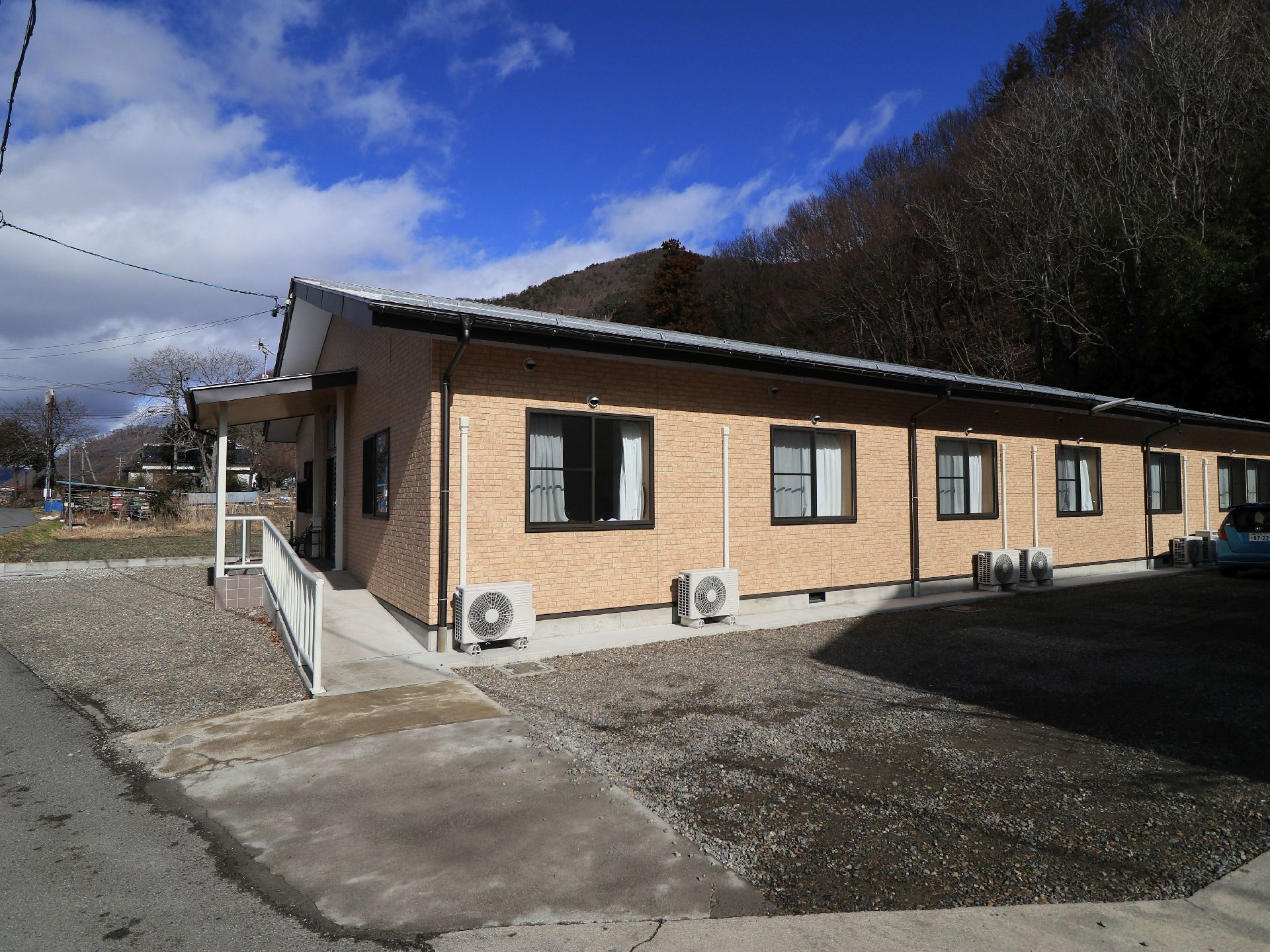 建物は白く、周囲に植物が植えられている。看板に施設名が書かれている。駐車場には屋根がついており、数台の車を止めることができる。