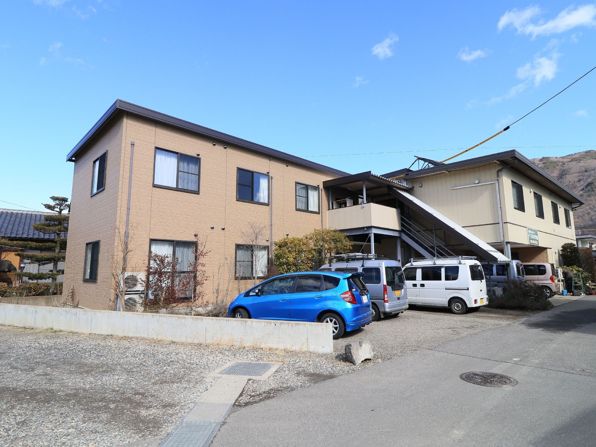 ベージュ系の２階建ての施設建物があり、手前に駐車場が設けられている。車が数台停められているのが見える。
