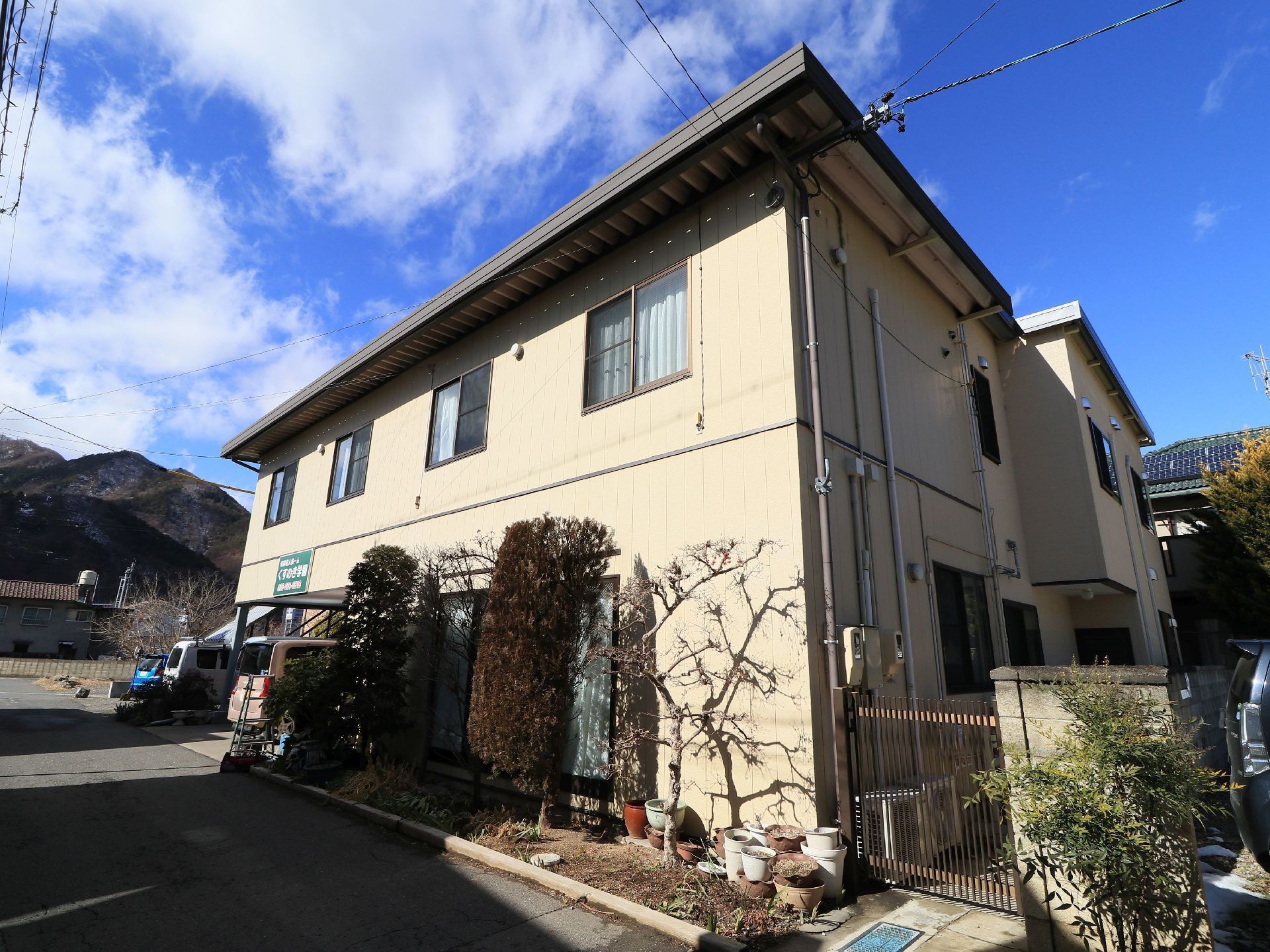晴れた日の山々を背景にした２階建て施設である。ベージュ系の建物で駐車場を完備し、一般住宅のような植え込みがある。