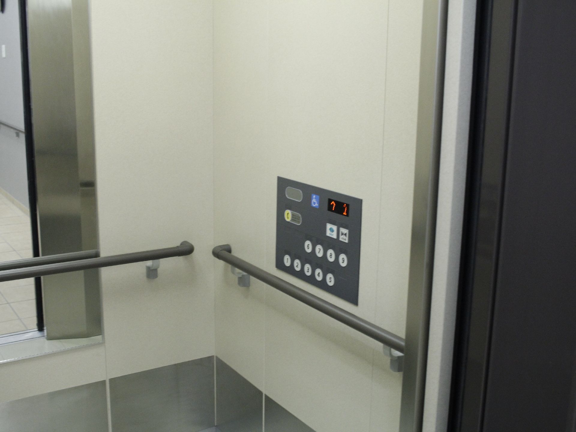 エレベータの内部の壁にはアイボリーとグレーが使われている。壁には手すりがあり、その上には操作パネルが設置されている。