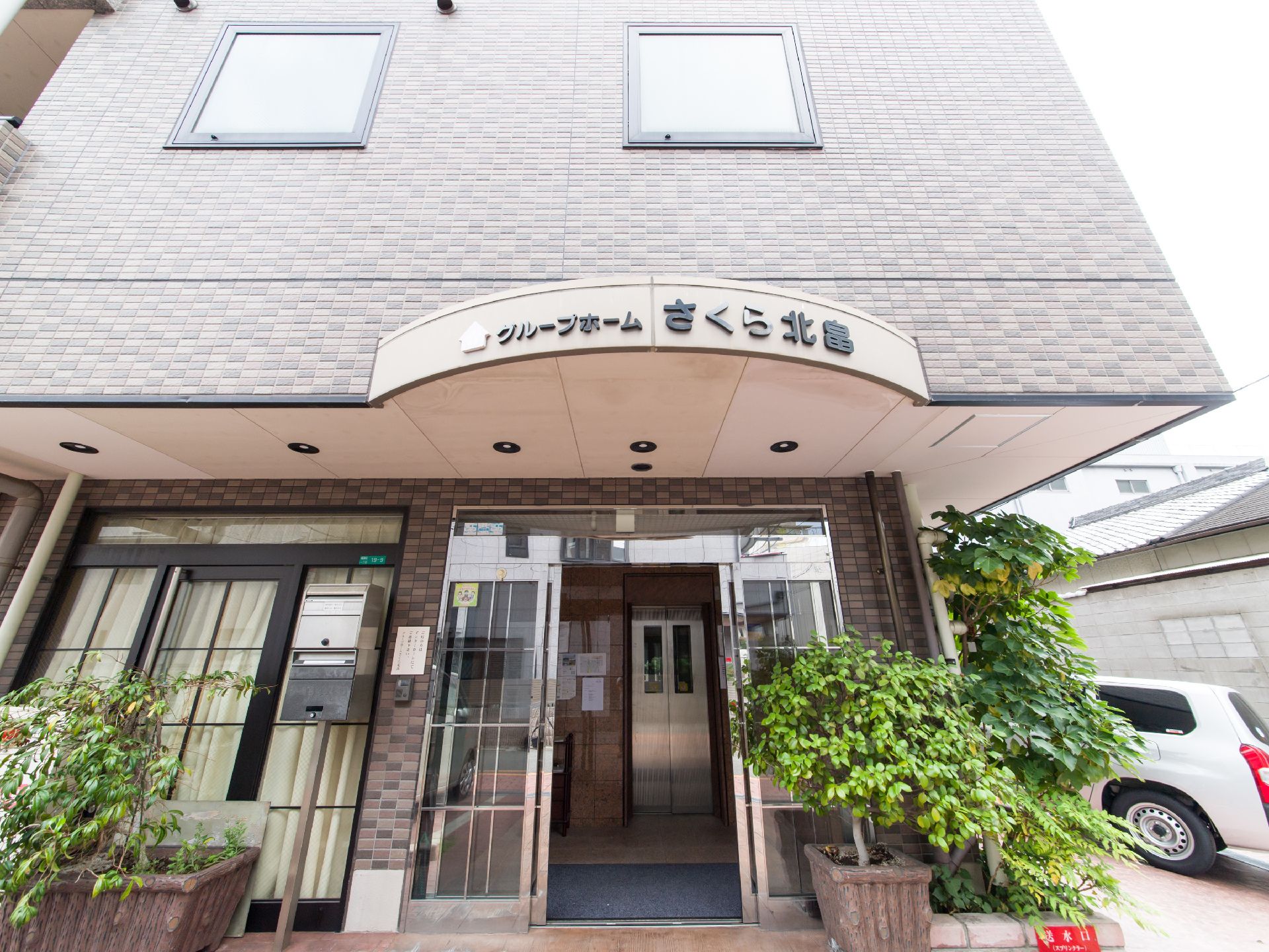 施設の玄関ドアの上にはベージュ系の屋根が設けられている。前面は曲線になっており、黒い文字で施設名が表示されている。