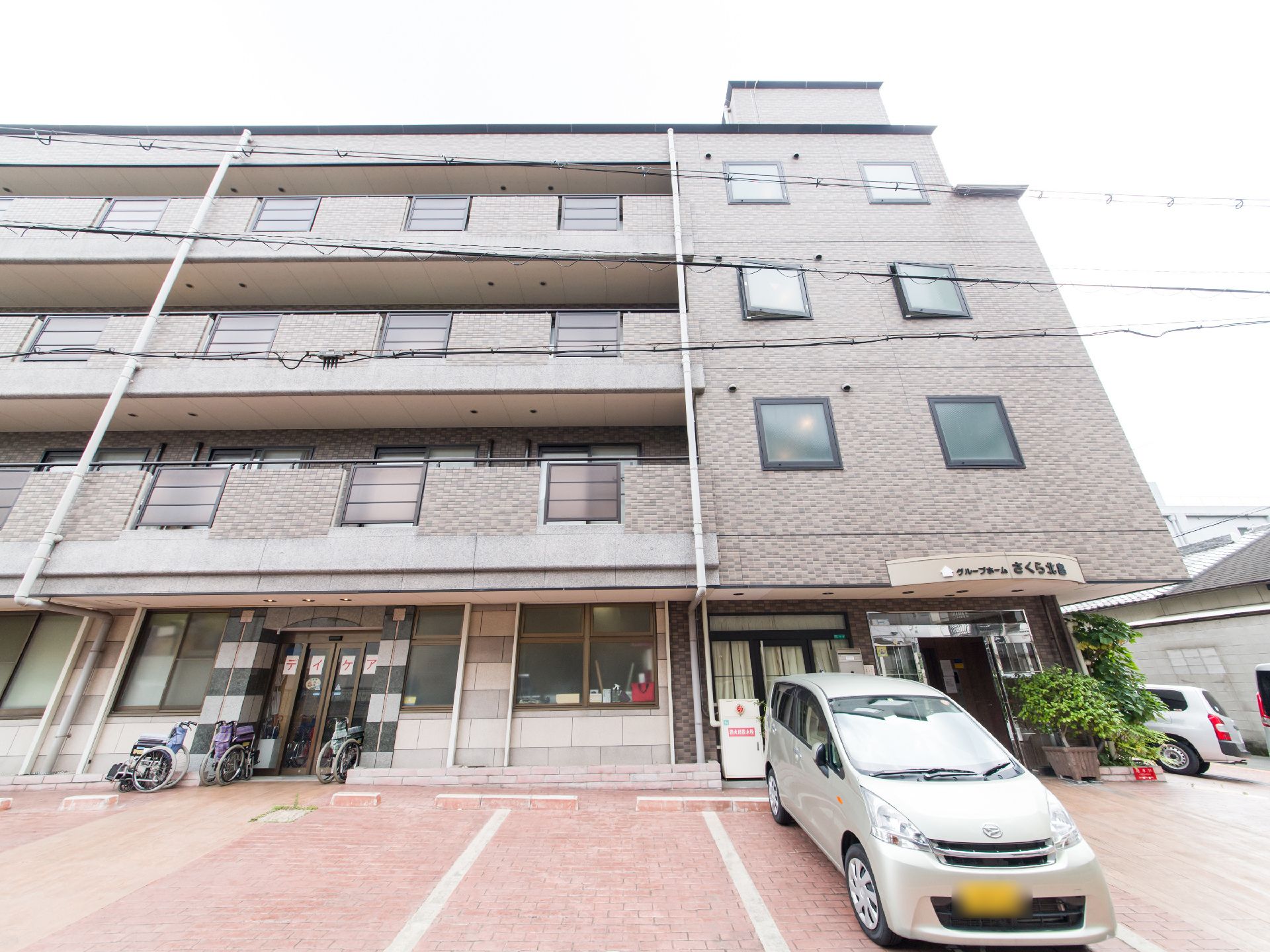 施設は4階建ての茶色い建物で、窓とフェンスのフレームは黒である。玄関がある右側部分は外開き窓が設置されている。