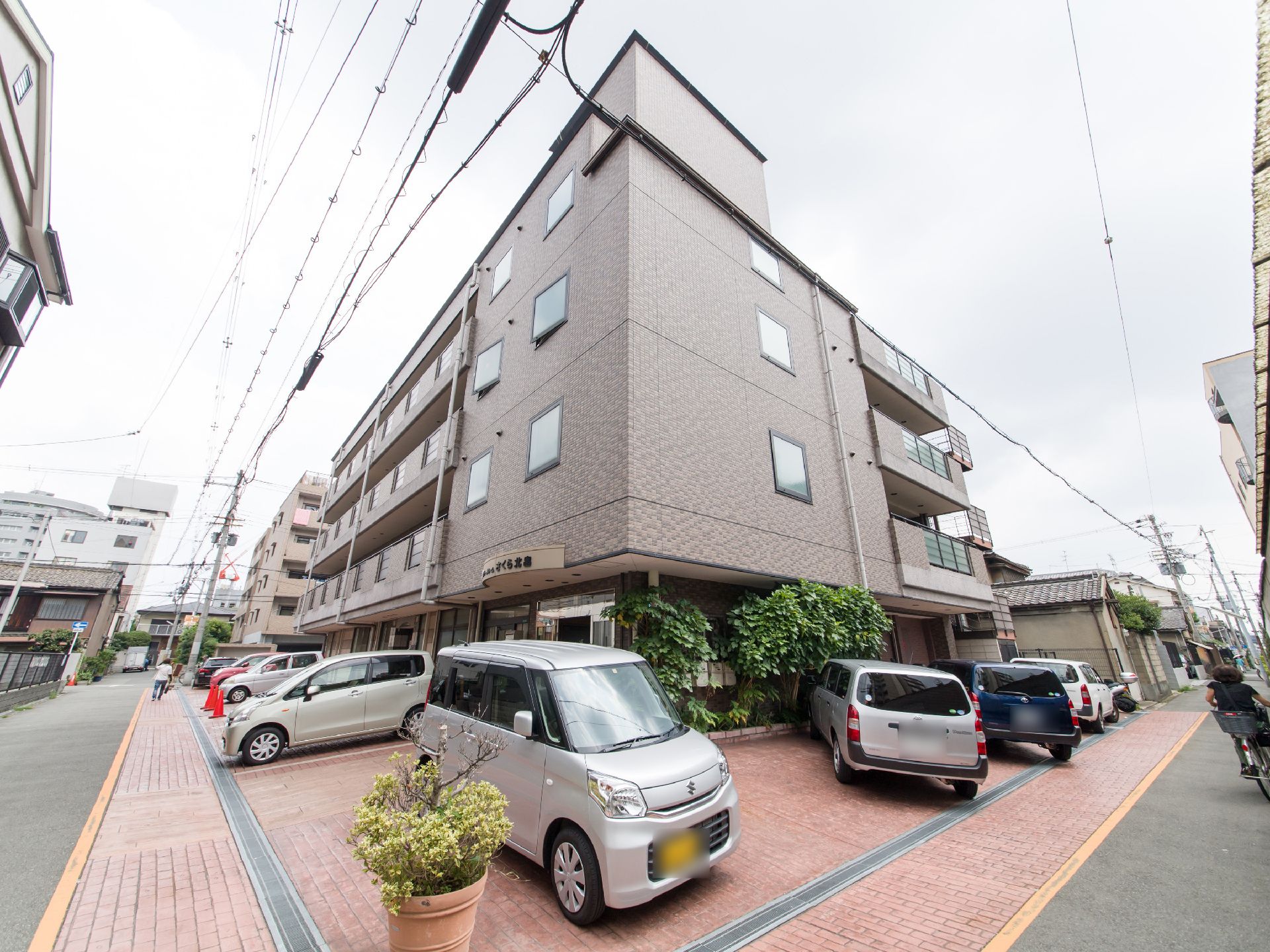 施設の建物は道路の曲がり角部分に建っている。玄関前と右側にはブラウン系のタイルが敷かれた駐車スペースがある。