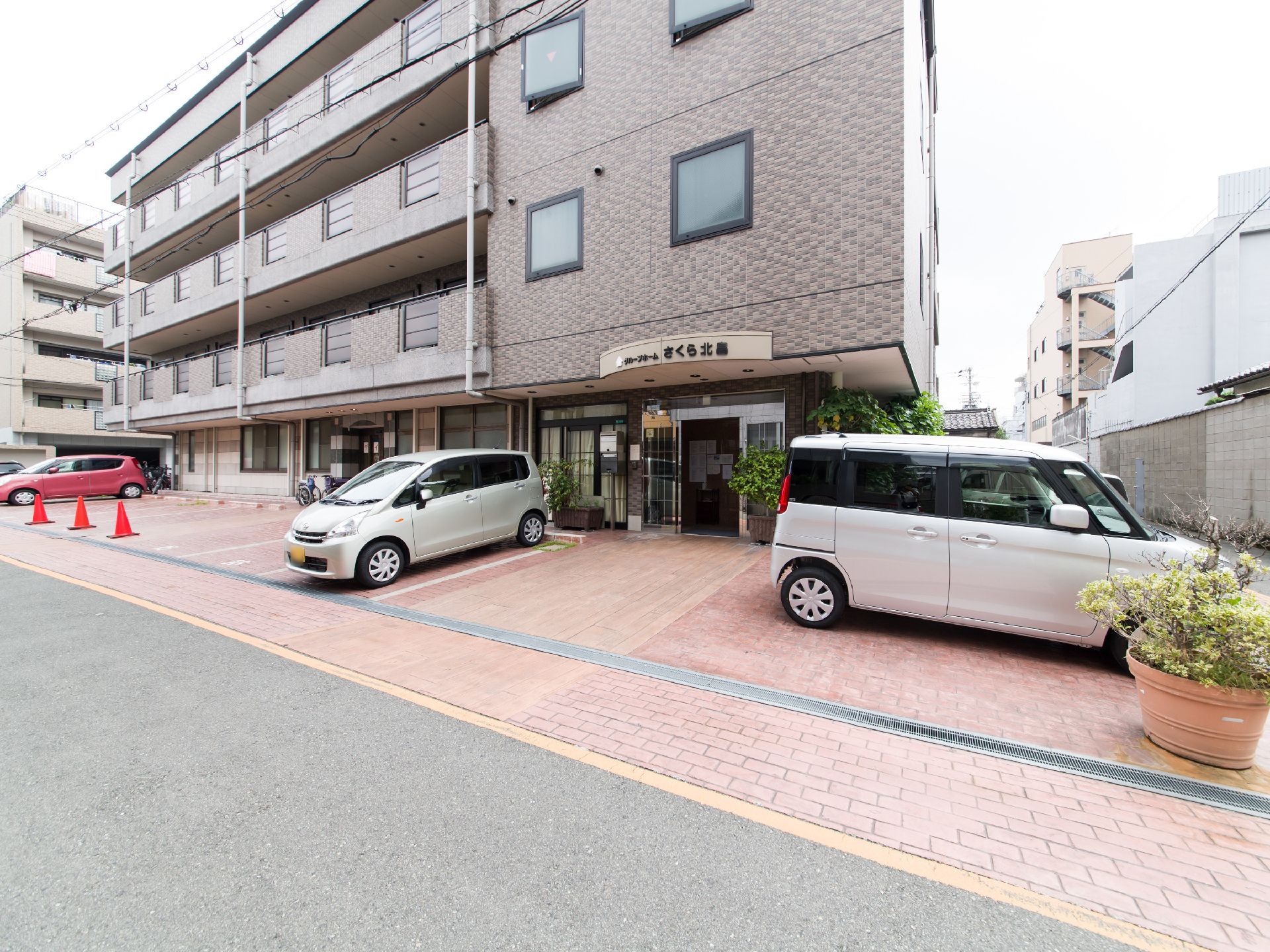 施設の建物の外壁はブラウン系のタイル張りである。建物前の床も同系色のタイル敷きで、駐車スペース用のラインが引かれている。