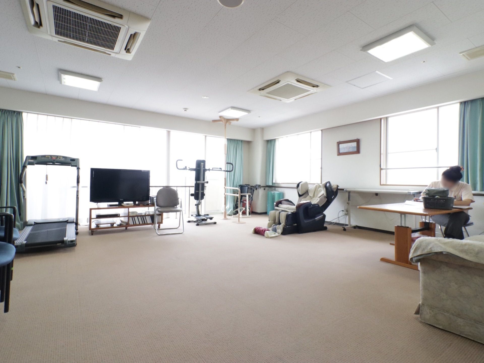広々とした部屋がある。大きな窓にはレースと薄い水色のカーテンがかけられている。テレビの横に機能訓練をする器具が置かれている。
