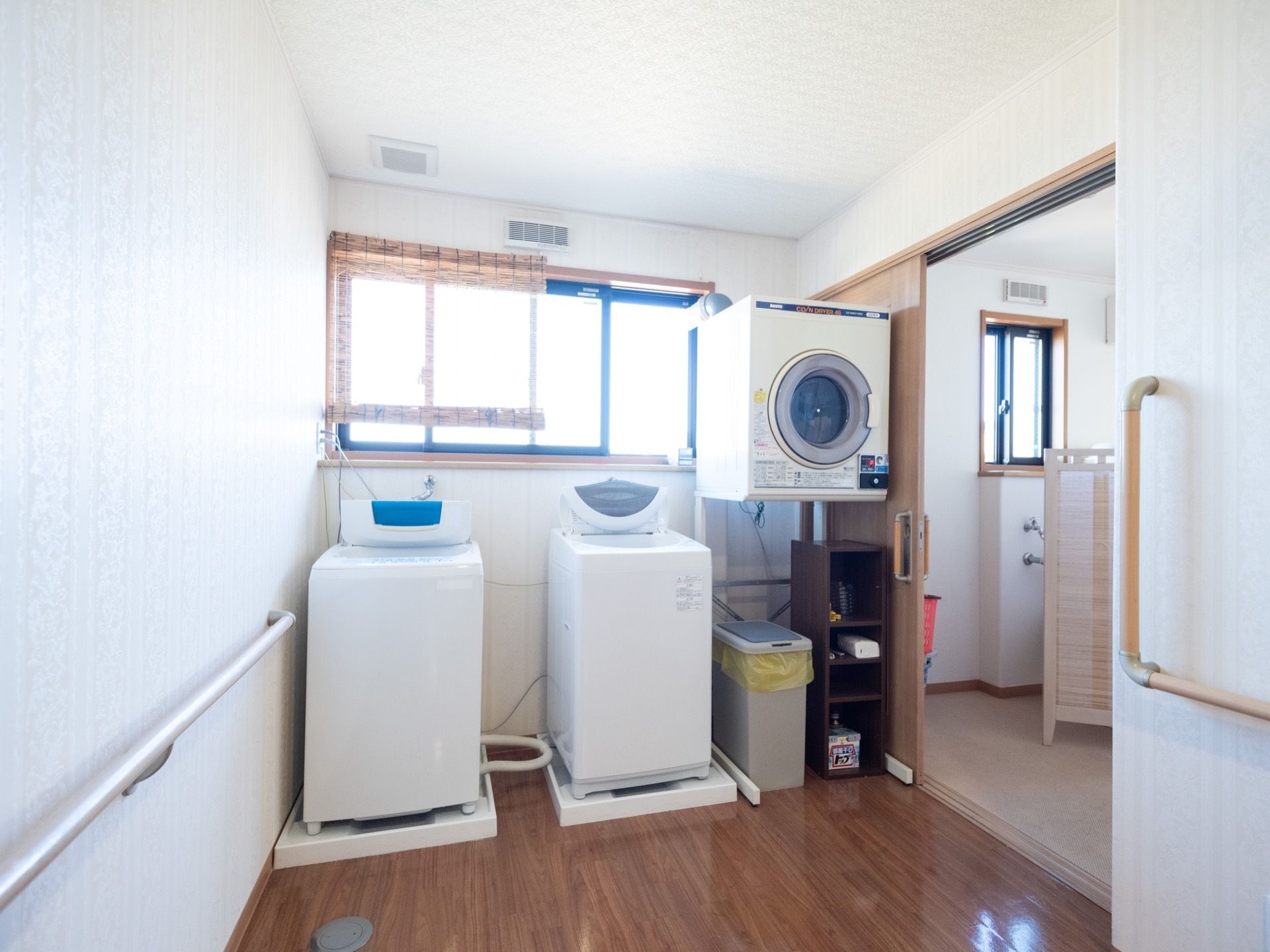 窓の前に簾が掛けられている。洗濯機が2台並べて設置されており、右側に乾燥機が置かれている。乾燥機の下にゴミ箱がある。