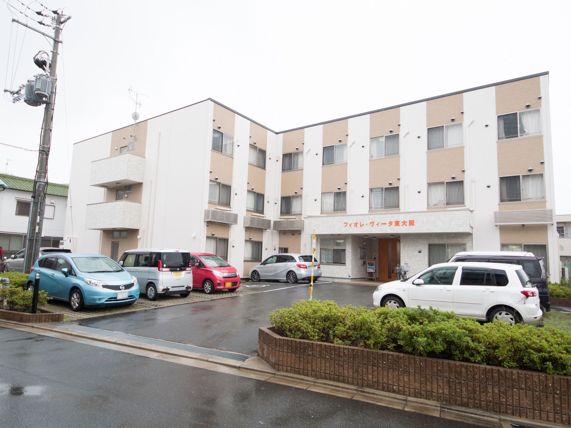 白とオレンジのモザイク状に塗られた３階建ての建物である。施設前は駐車場があり、たくさんの車が停まっている。
