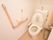 白色を基調とした室内には、タンク付きのトイレが設置されている。壁には、手すりとトイレットペーパー掛けが取りつけられている。