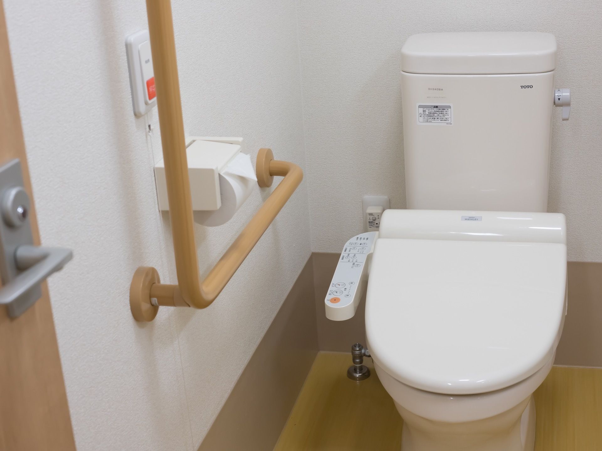温水洗浄機能付きの白く清潔感のあるトイレである。壁にはＬ字型の手すりや、緊急呼び出しボタンが備えてある。