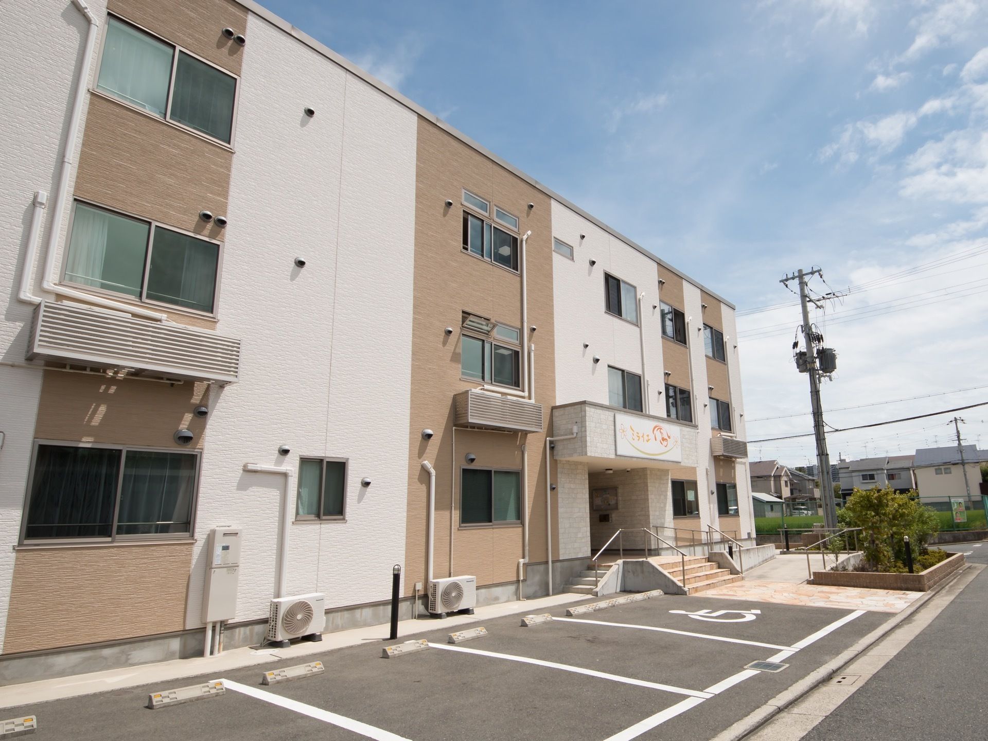 オフホワイトとブラウンのツートンカラーの建物である。建物のすぐそばに駐車スペースがある。各部屋の窓に網戸が設置されている。