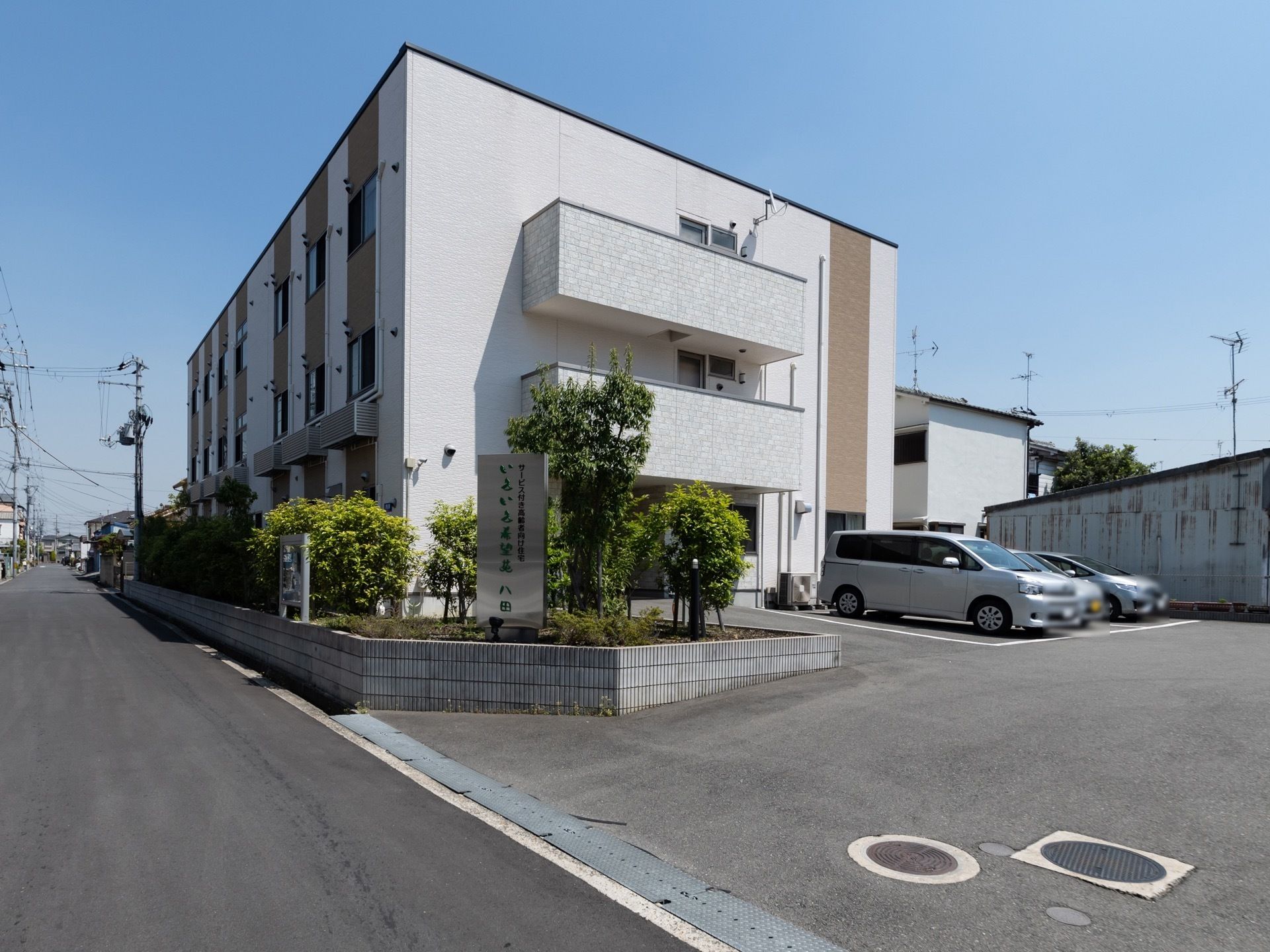 敷地内と道路の境目に低めの塀を立てていて、木などを植えている。入り口にはシルバー色の看板が設置されている。